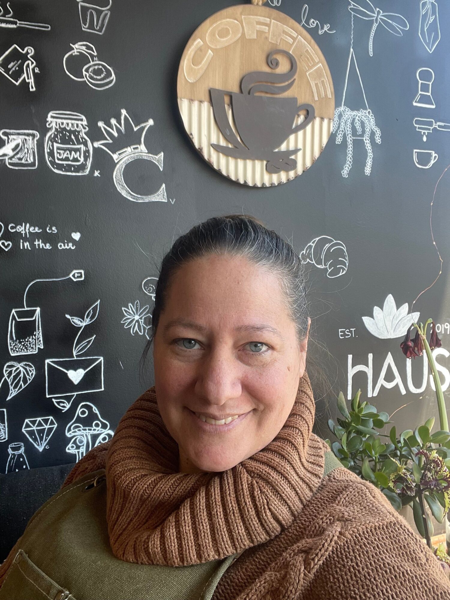 Smiling woman with dark hair tied back, wearing a brown knitted scarf, in front of a chalkboard with coffee-themed drawings.
