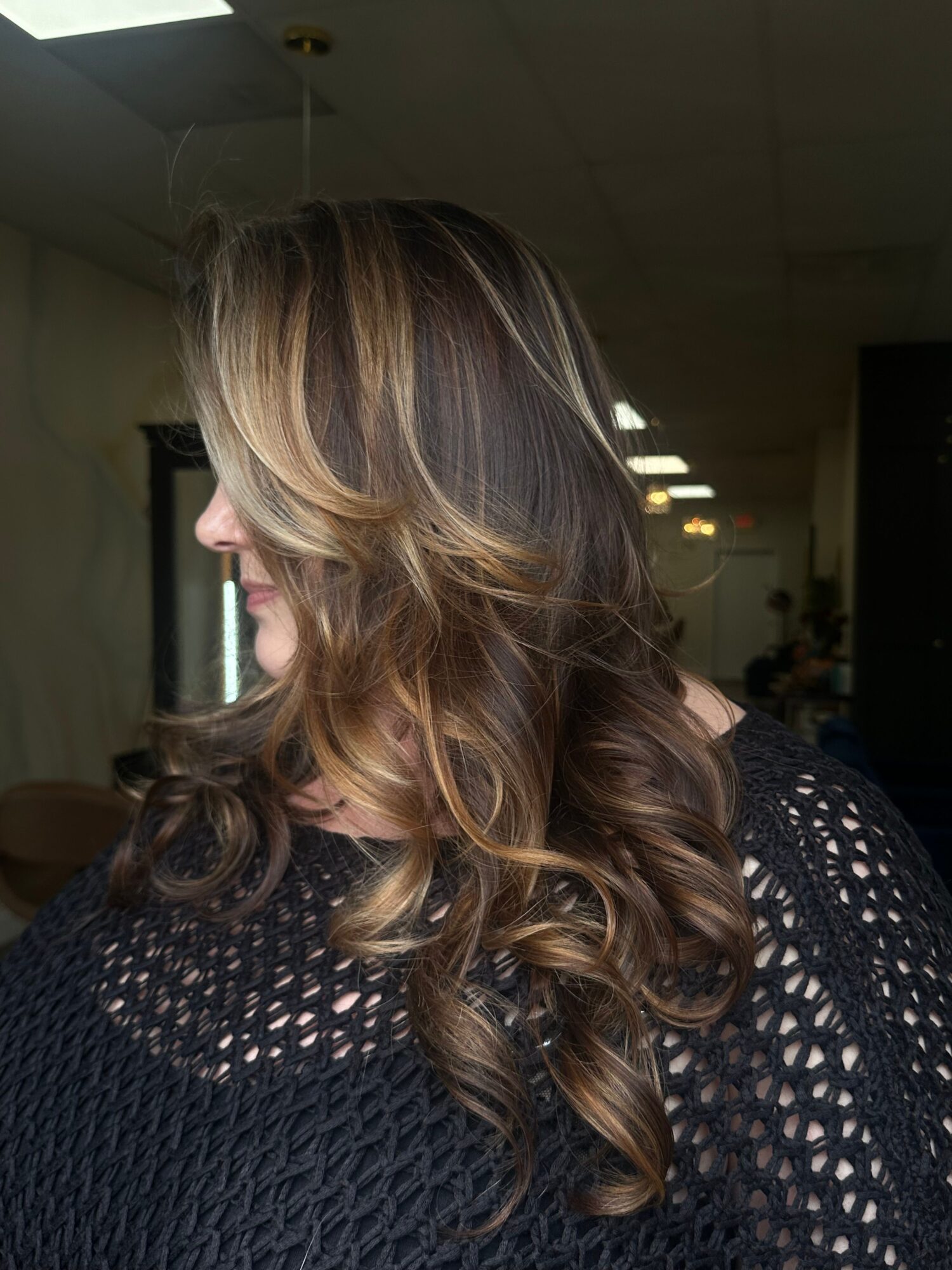 Side view of a woman with long, wavy hair and a black, open-knit top indoors.