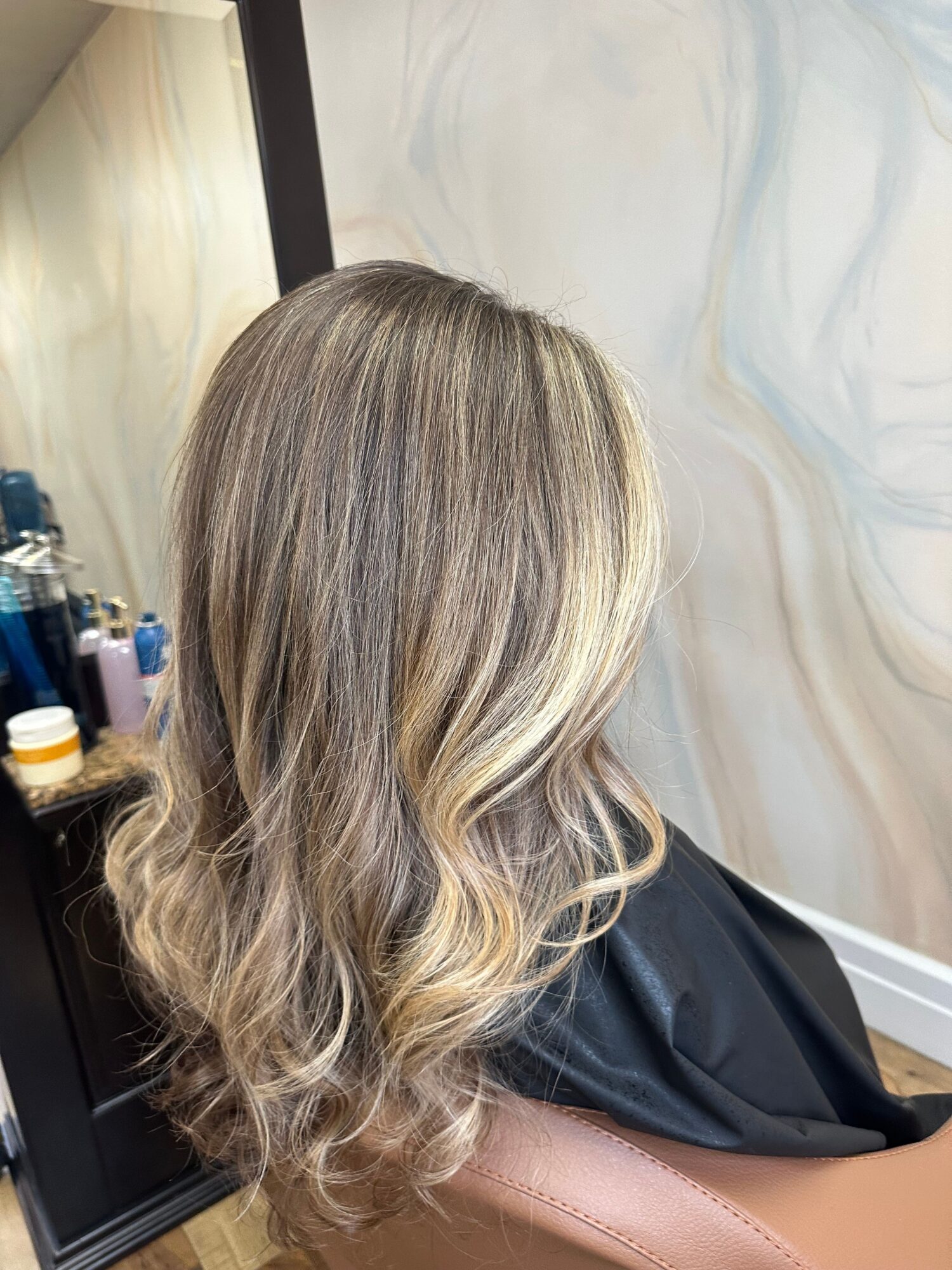 Back view of a woman with long, wavy, blonde hair sitting in a salon chair.