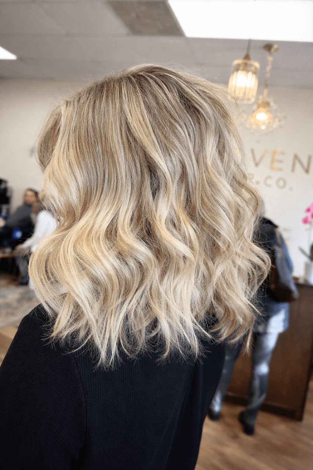 Person with shoulder-length wavy blonde hair in an indoor setting with hanging lights and people in the background.