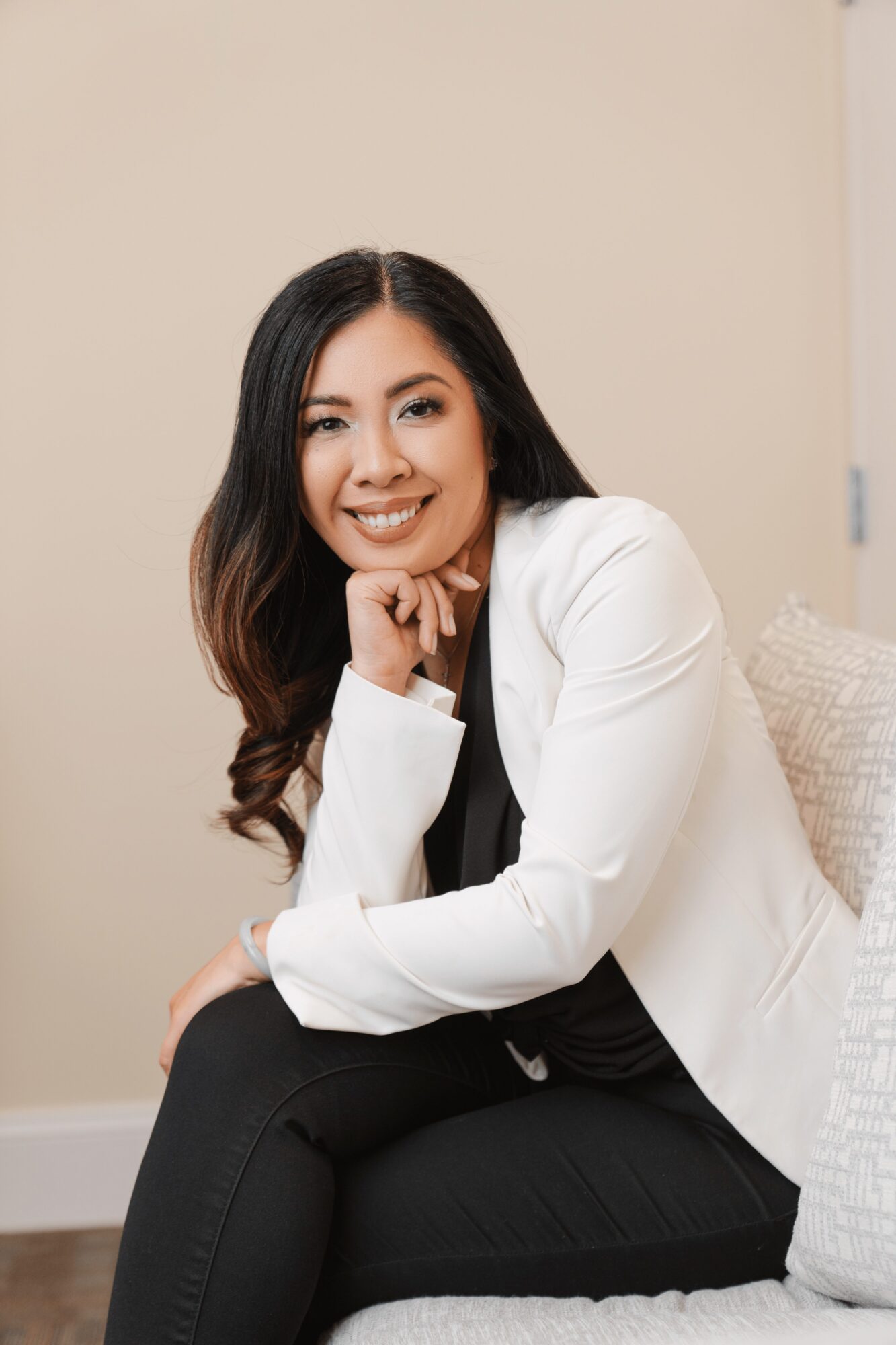 Woman with long dark hair smiling, sitting on a couch with a beige wall background.