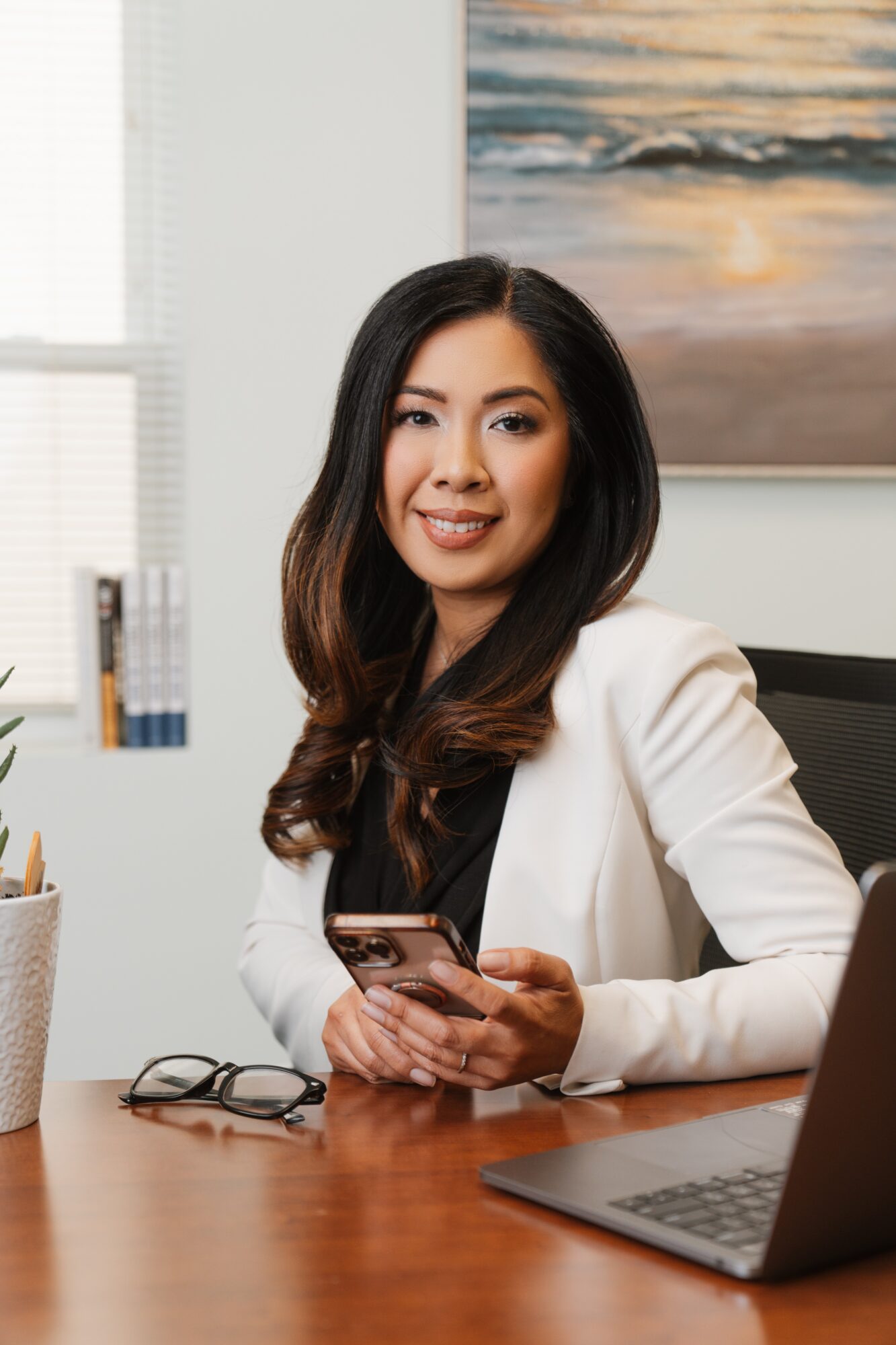 Woman with long dark hair in white blazer sitting at desk holding smartphone, office background with painting.