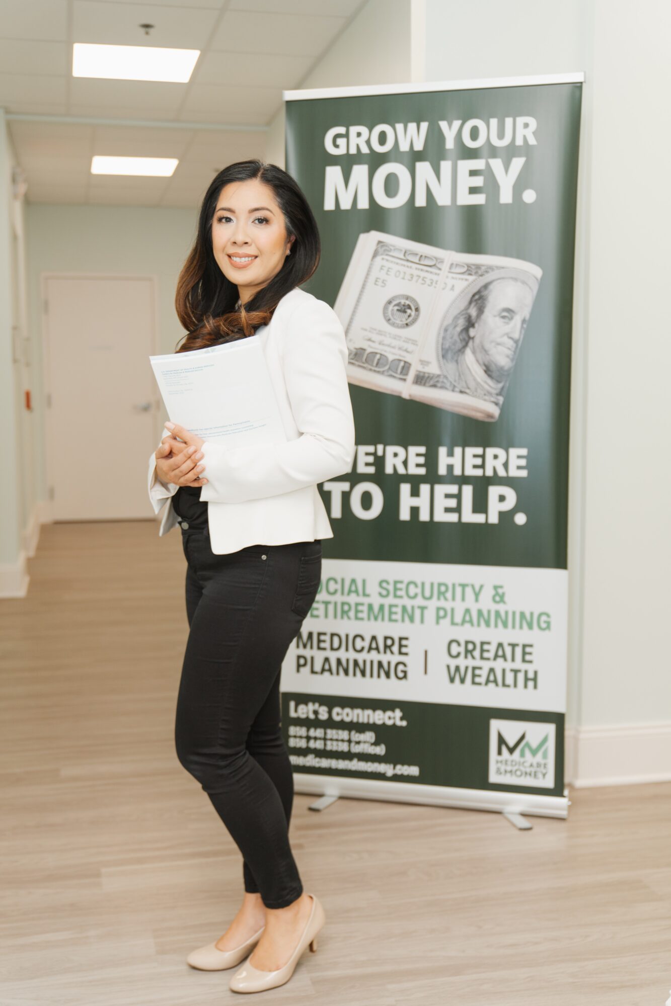 Woman in white blazer and black pants standing next to a green financial planning banner, smiling, holding papers.