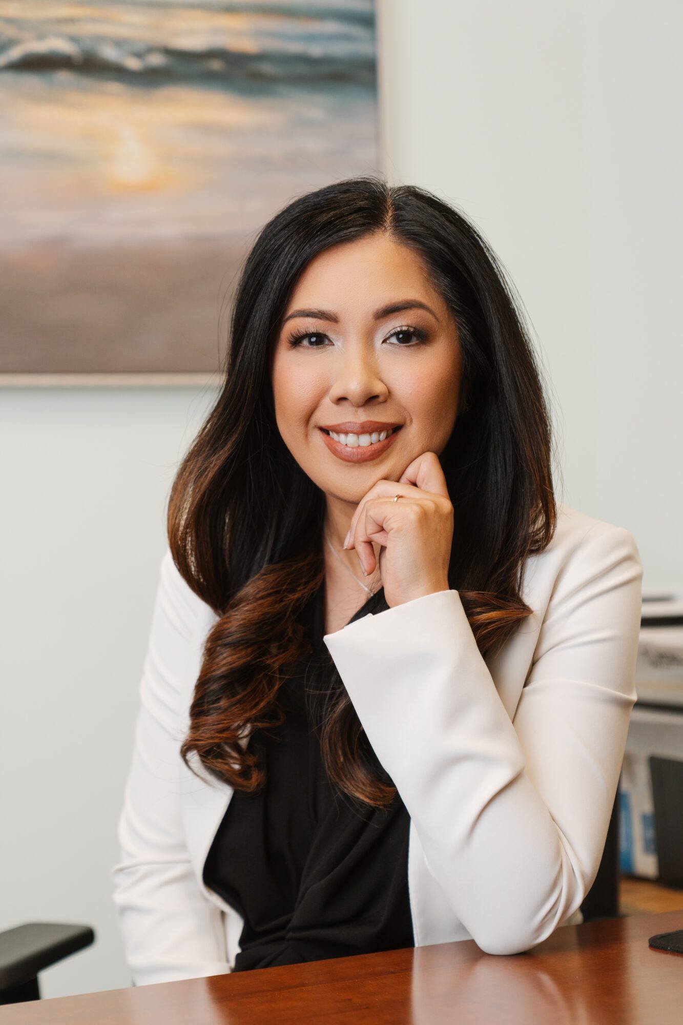 Young woman with long dark hair smiling, sitting at a desk with a painting in the background.