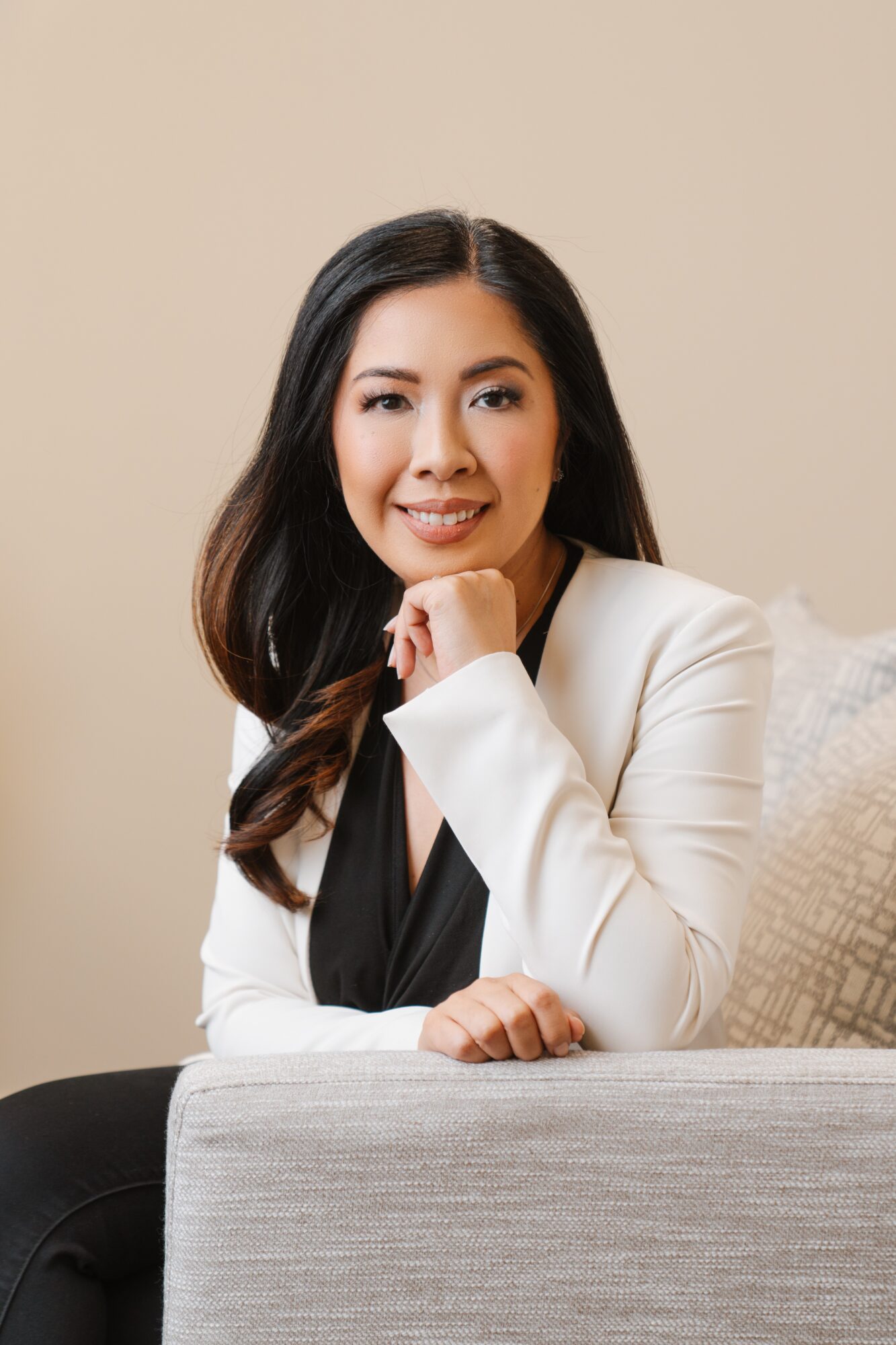 Woman with long dark hair smiling, resting her chin on her hand, sitting on a sofa in a white blazer.