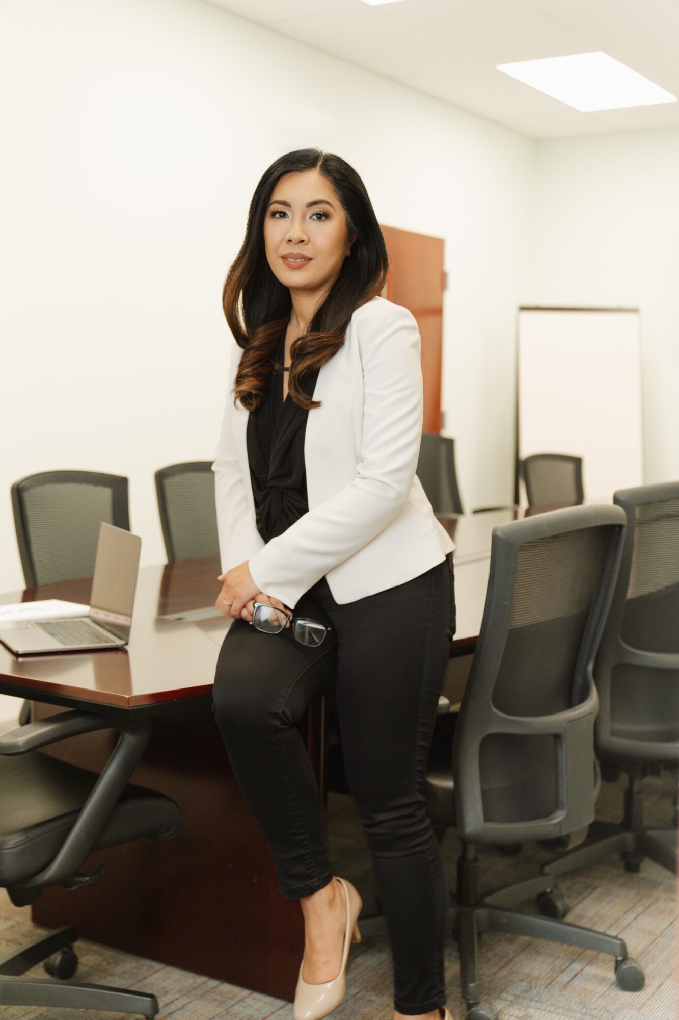 Woman standing in a conference room with chairs, a laptop, and a whiteboard behind her.