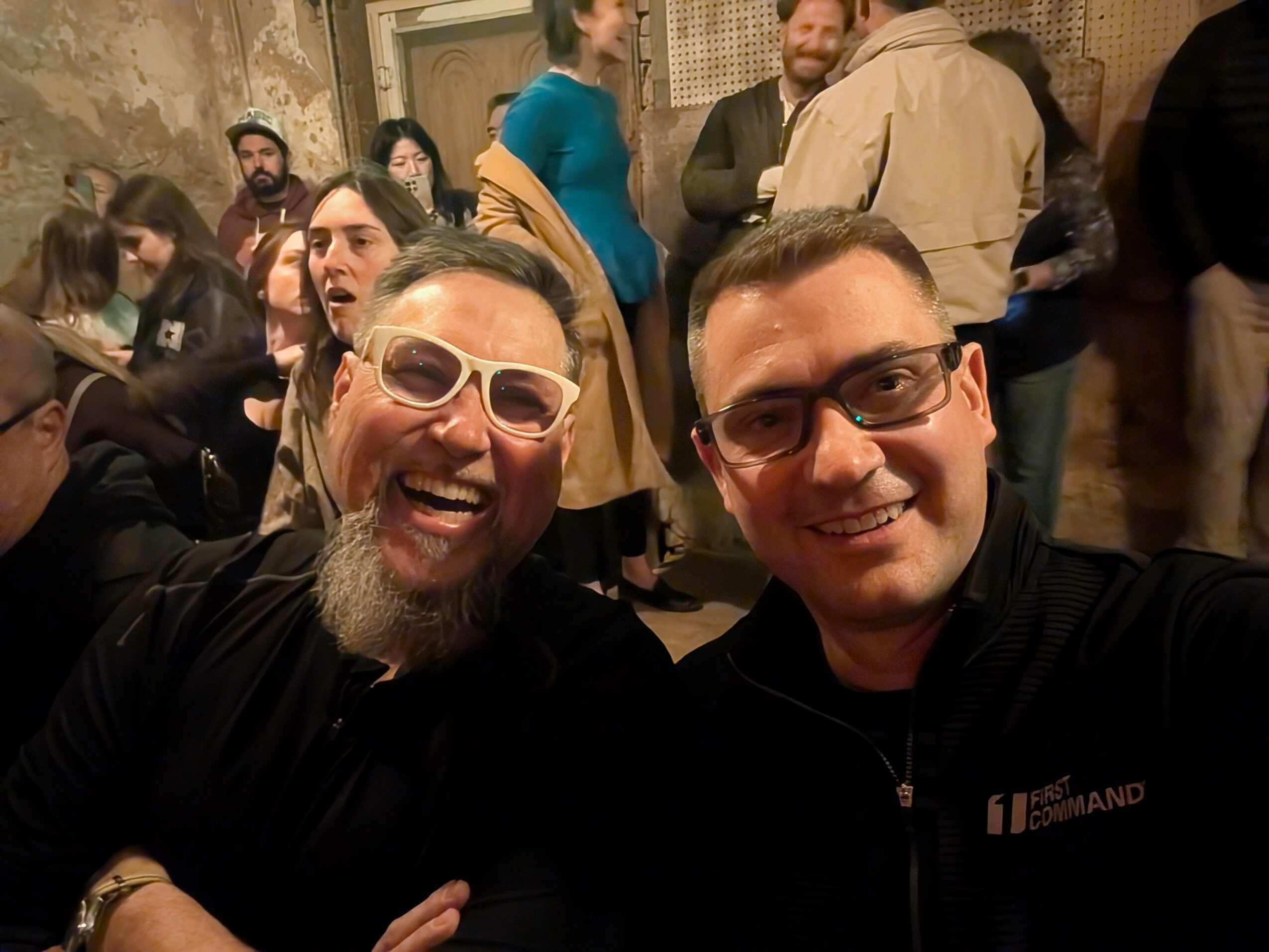 Two men smiling and taking a selfie at a crowded indoor event.