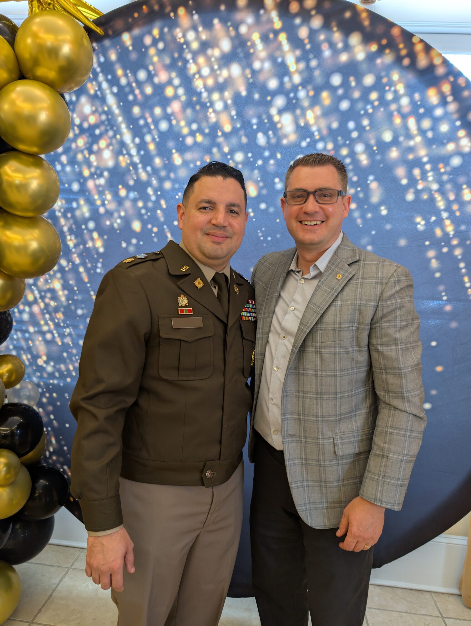 Two men standing together, one in military uniform and the other in a gray plaid blazer, smiling in front of a festive backdrop.