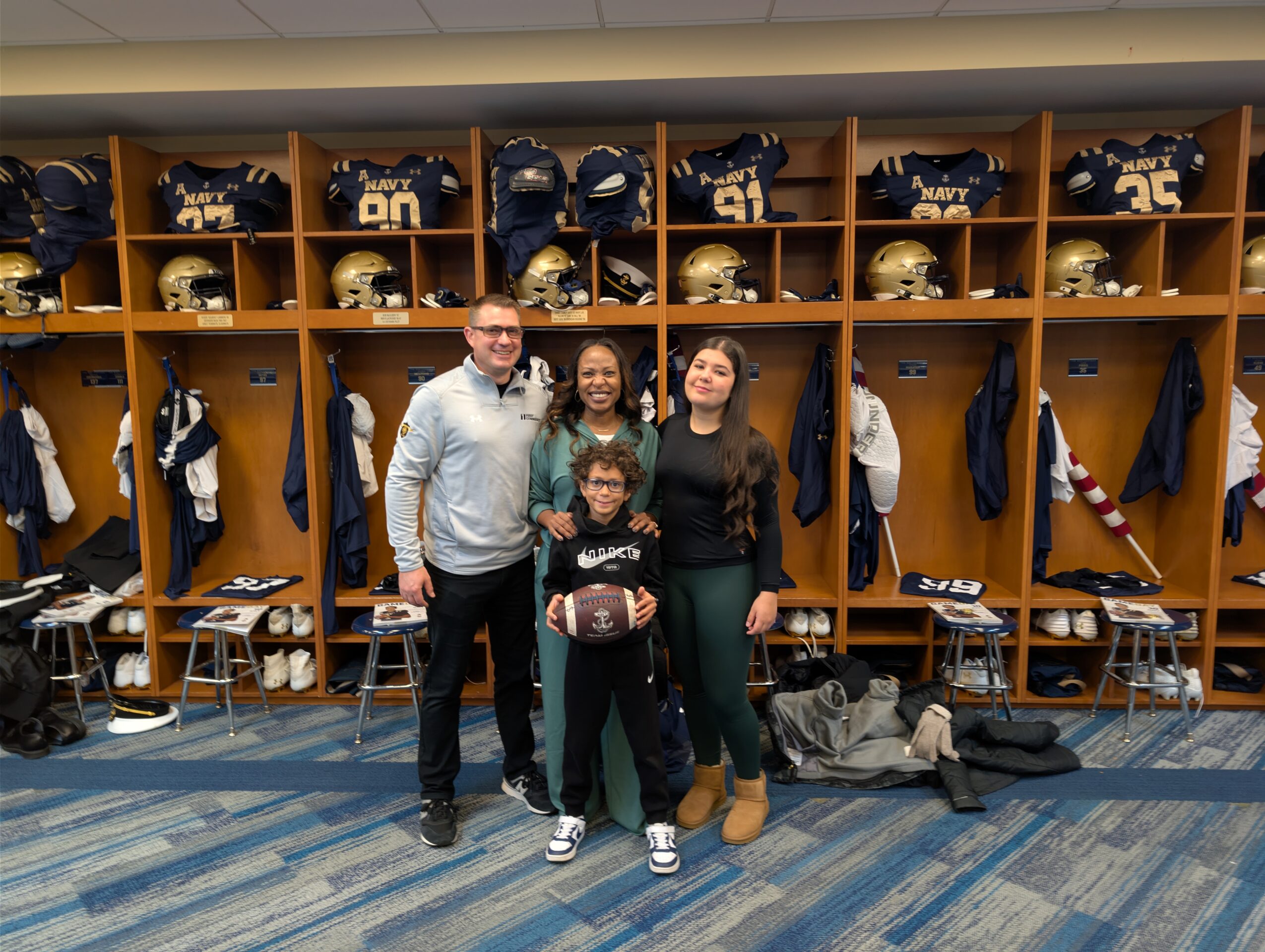 Four people stand in front of a football locker room with helmets and jerseys on shelves behind them.