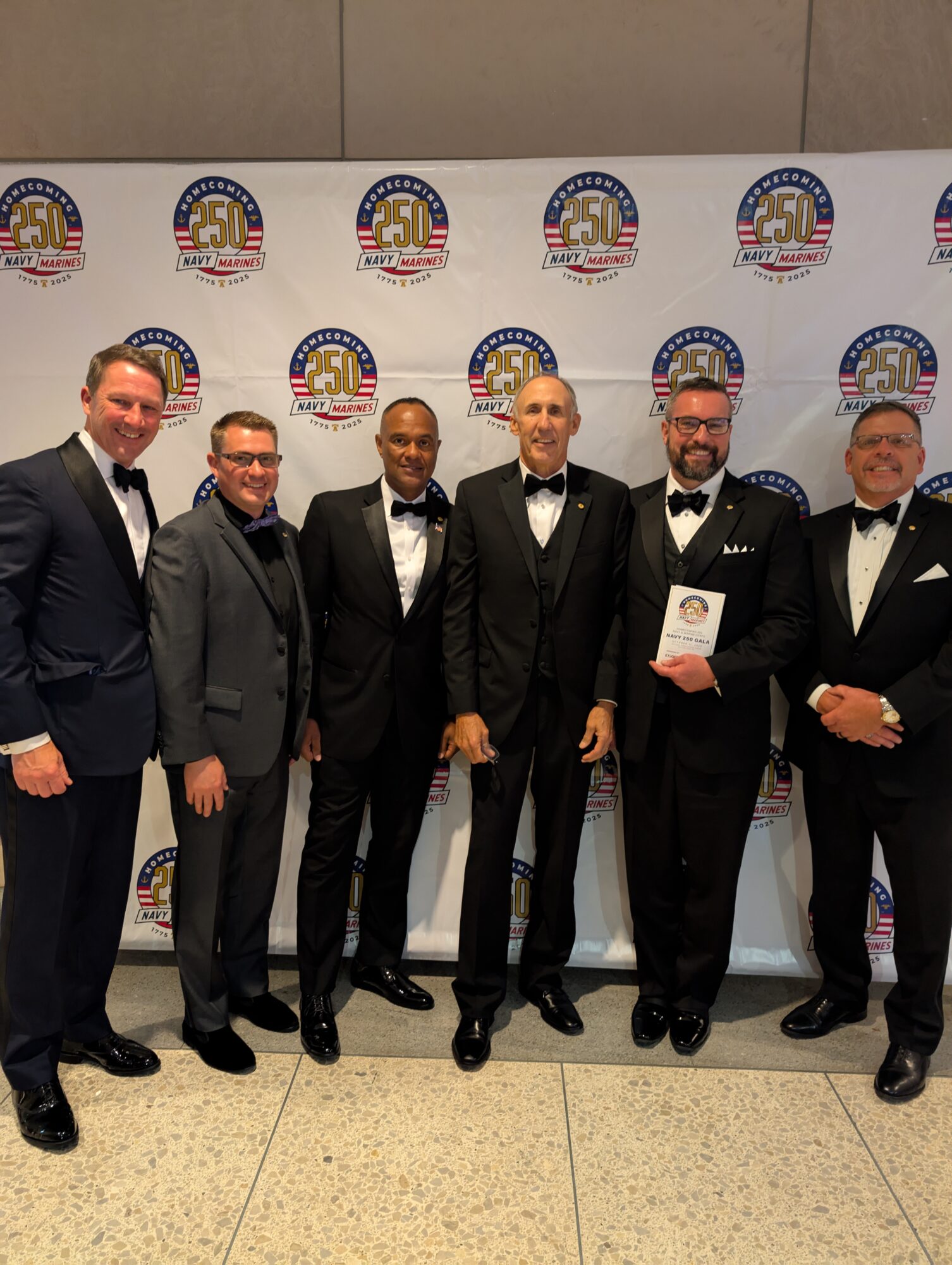 Six men in formal tuxedos standing in front of a backdrop with a logo, smiling for the photo.