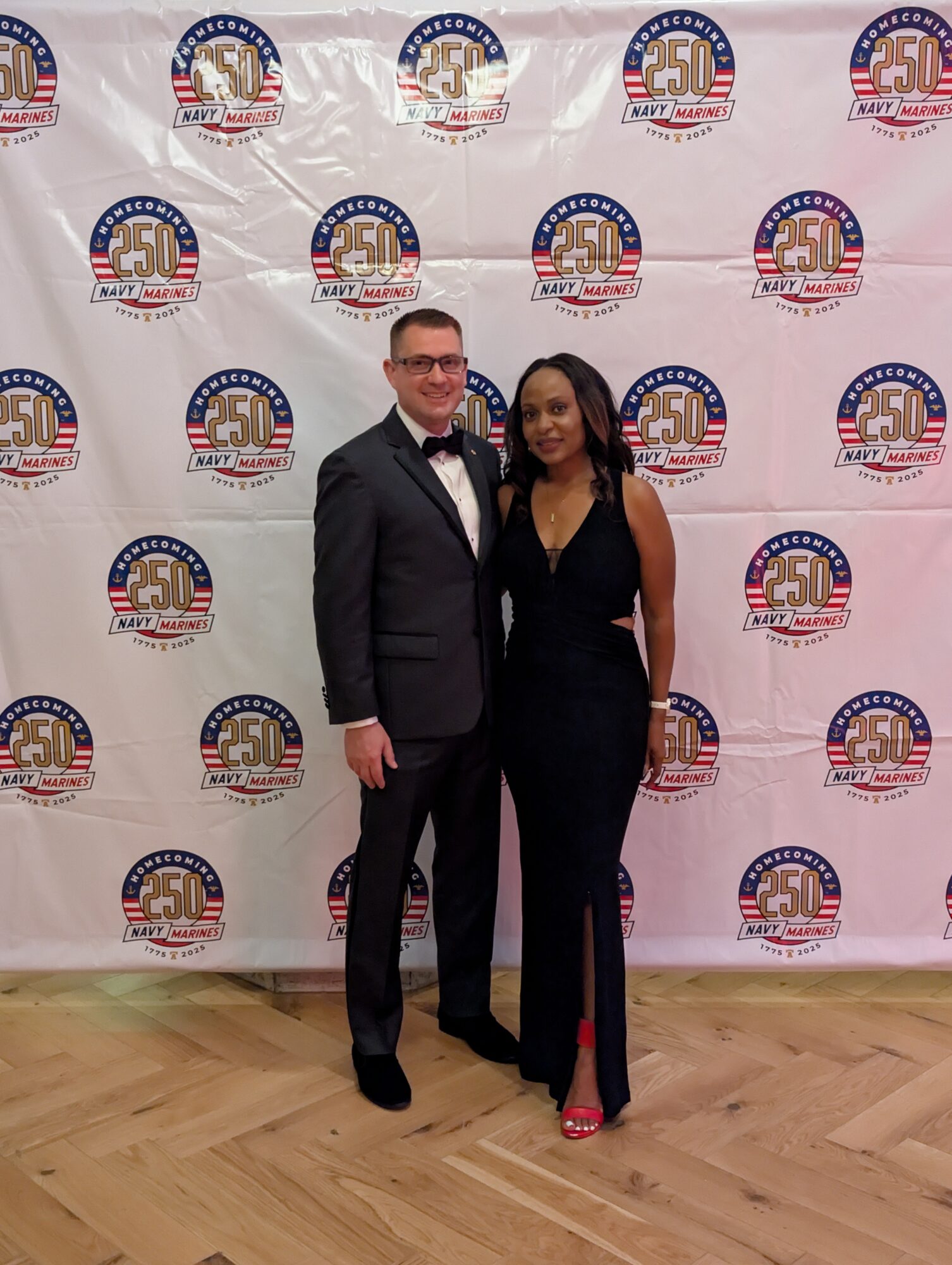 Man and woman in formal attire standing on wooden floor in front of a backdrop with American flag and 50th anniversary logos.
