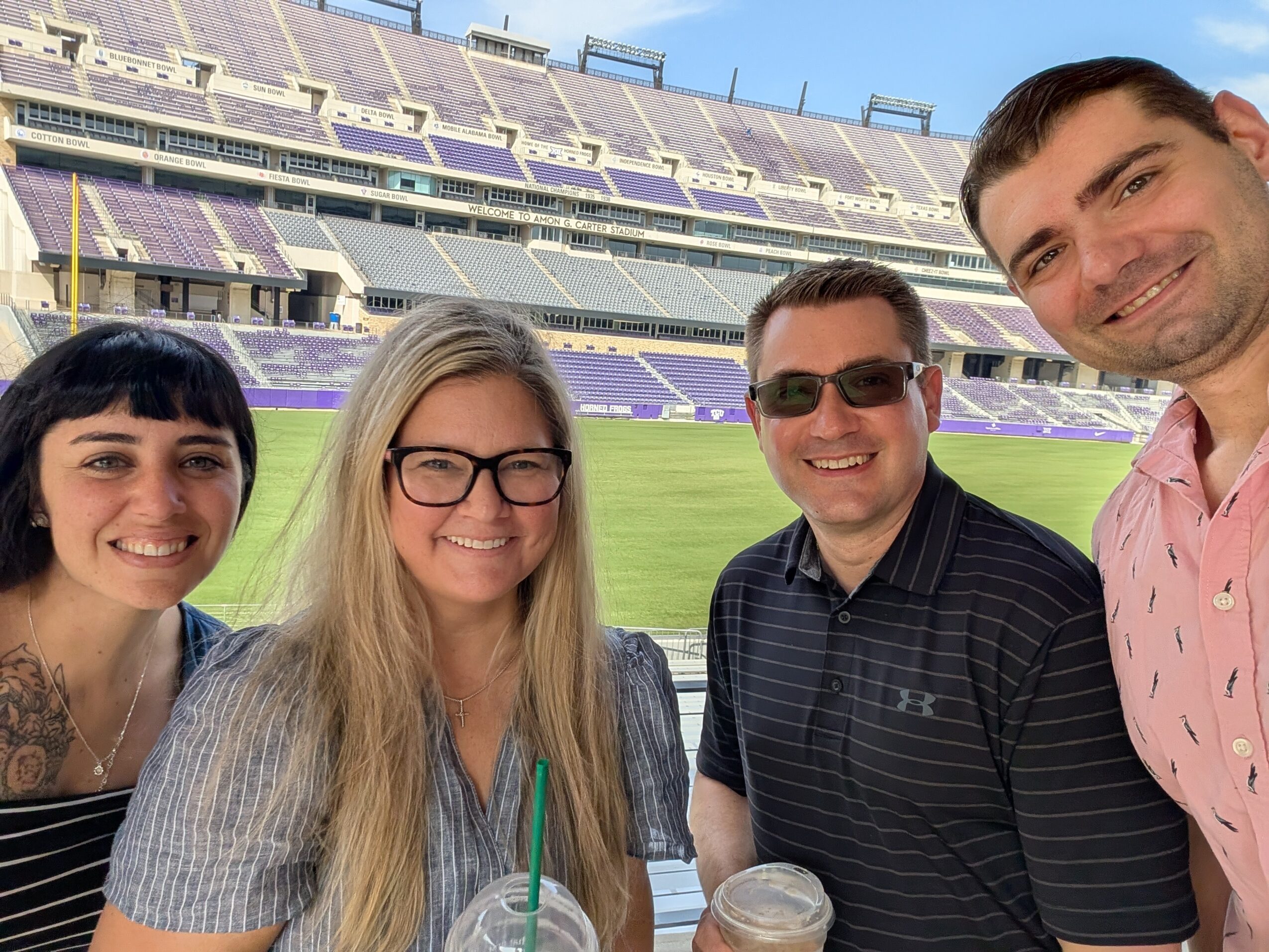Four people smiling at a stadium with empty seats and a field in the background.