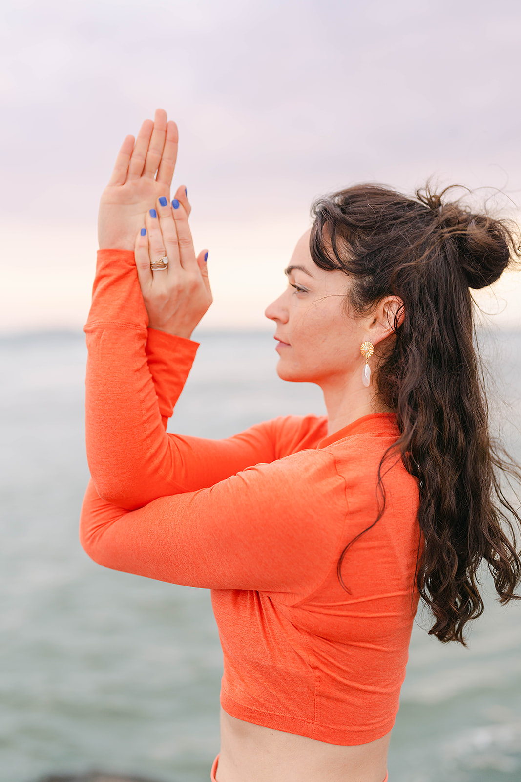 Woman with long dark hair in an orange top practicing yoga outdoors near water, with hands in prayer position.