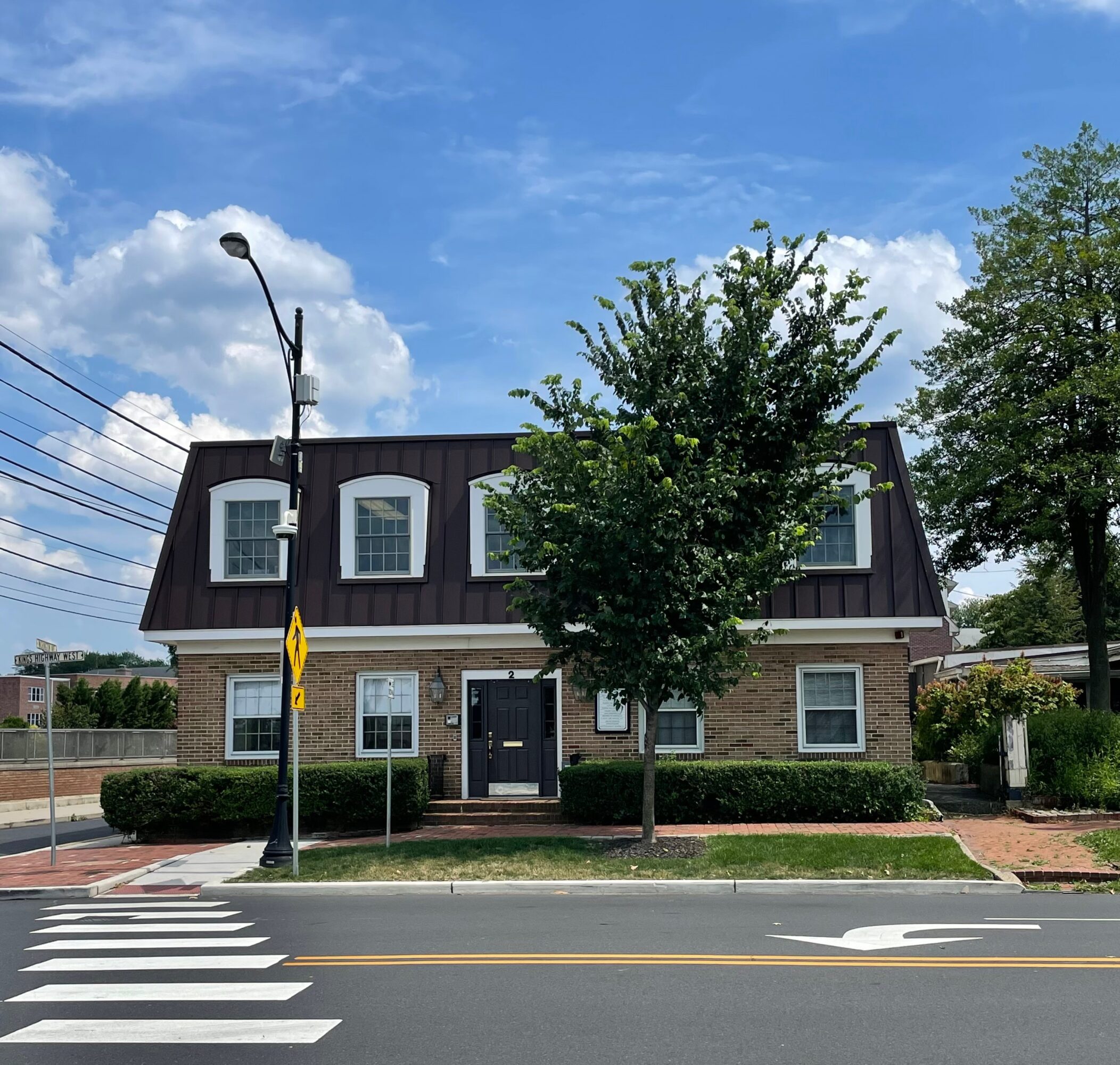 Two-story building with a tree in front, street, and crosswalk, under a blue sky with clouds.