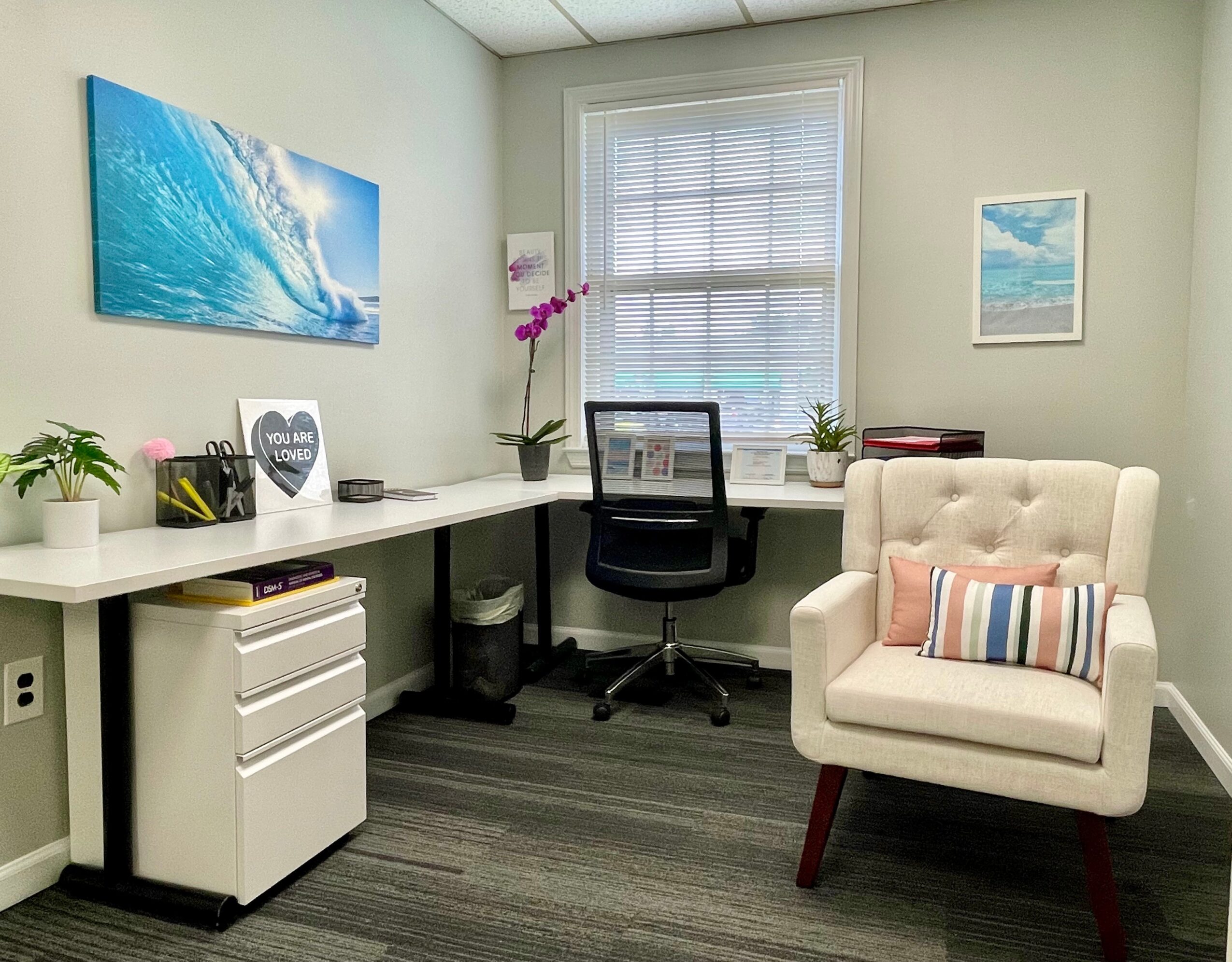 Office with desk, chair, armchair, window, and wall art, decorated with plants and a flower.