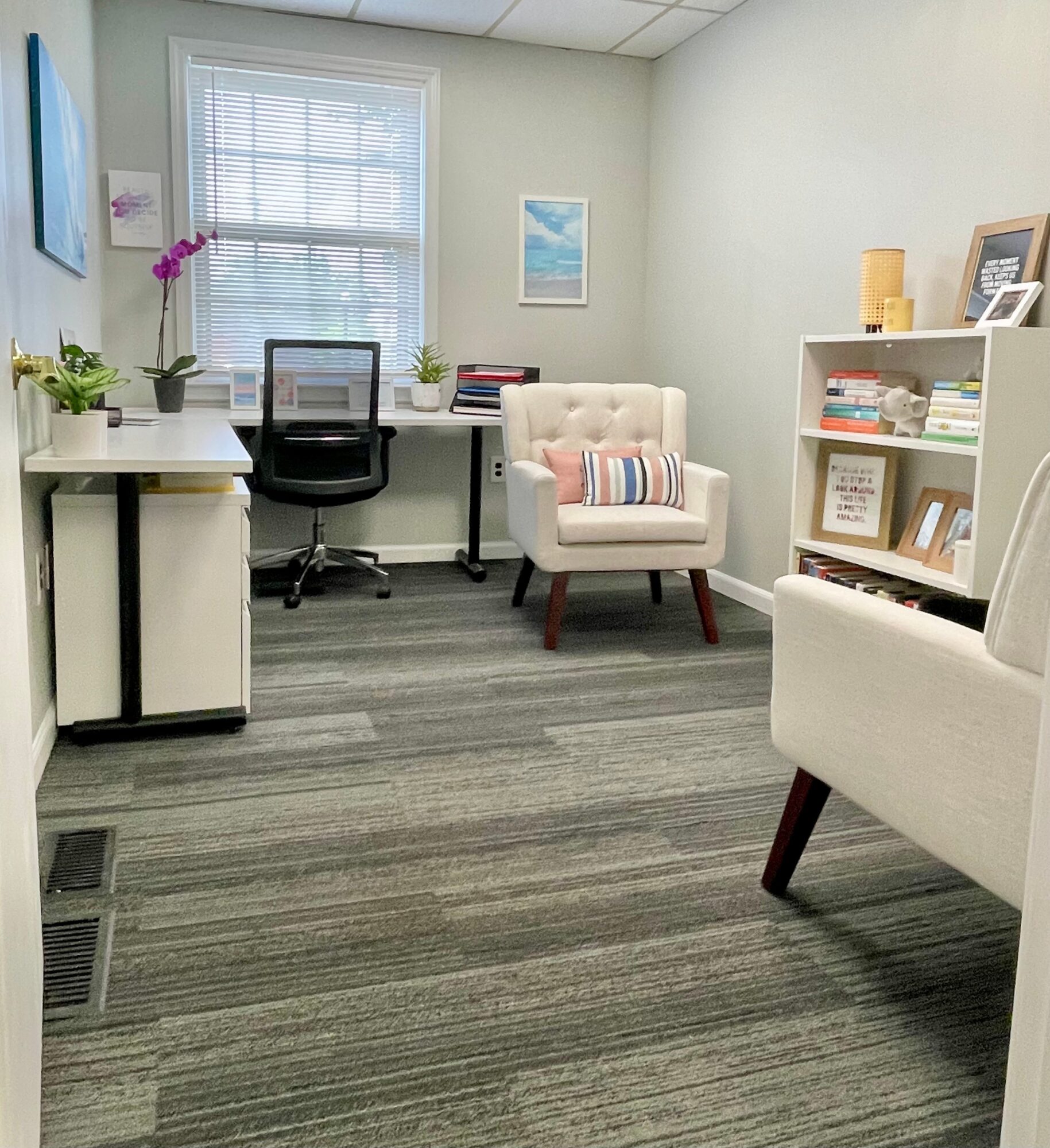Office room with desk, chair, white armchair, bookshelf, and window with blinds.