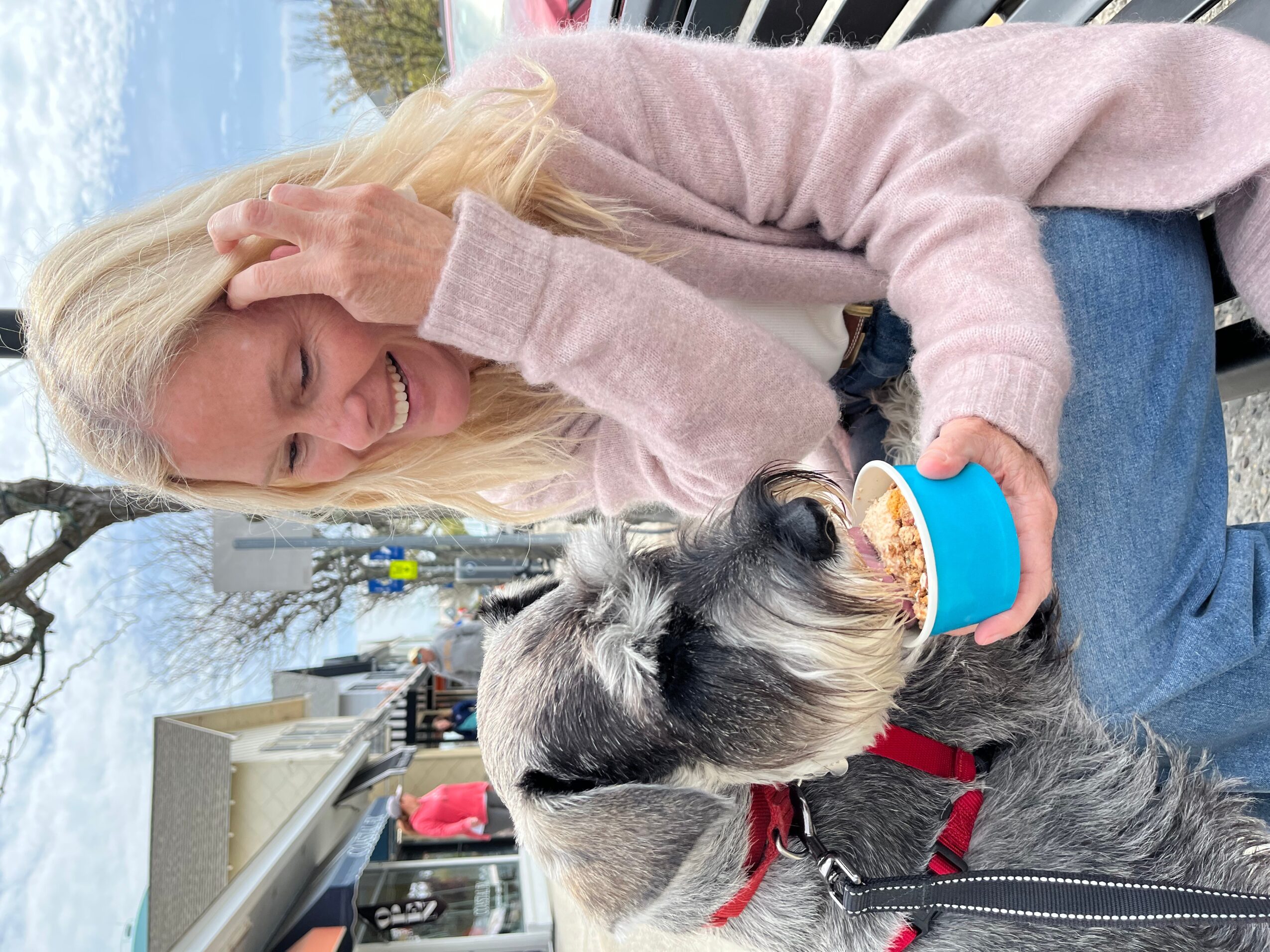 Woman with blonde hair smiling, holding a cup, sitting outdoors with a dog wearing a red collar.
