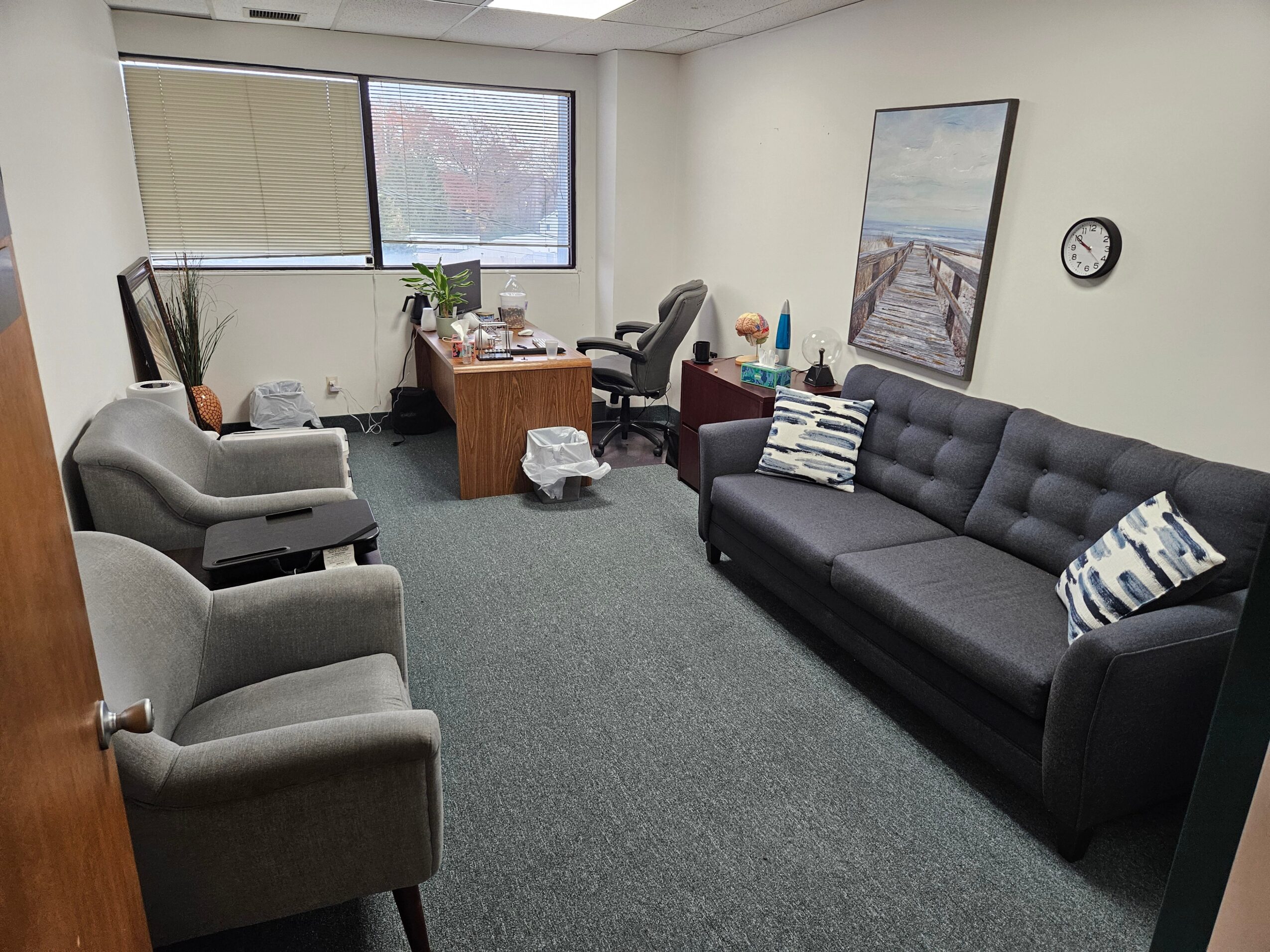 Office room with desk, chairs, sofa, window, clock, and various items on desk and sofa.