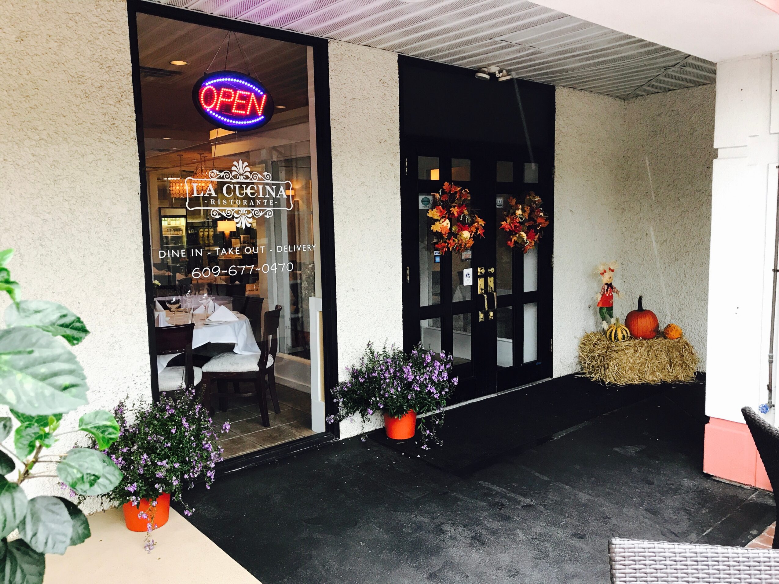 Restaurant entrance with flowers, pumpkins, hay bale, and an open sign, seen through a glass door and window.