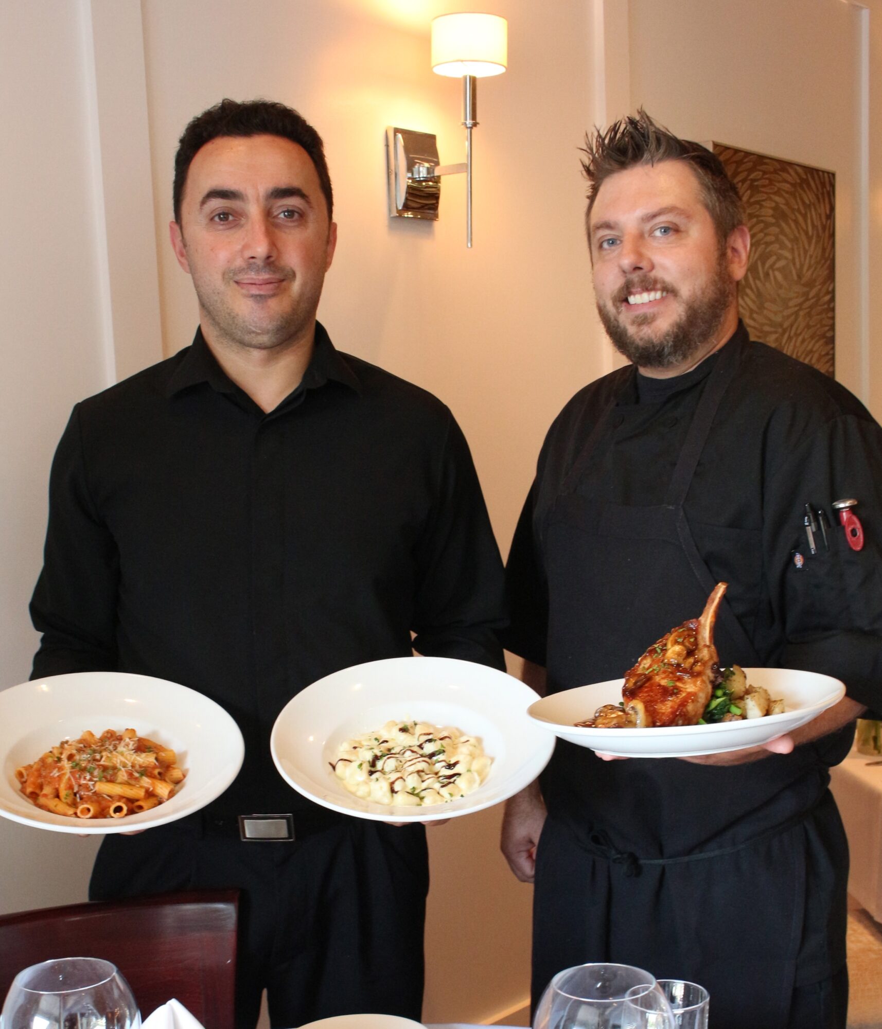 Two men holding plates of food, one in black shirt, the other in chef's uniform, in a restaurant setting.