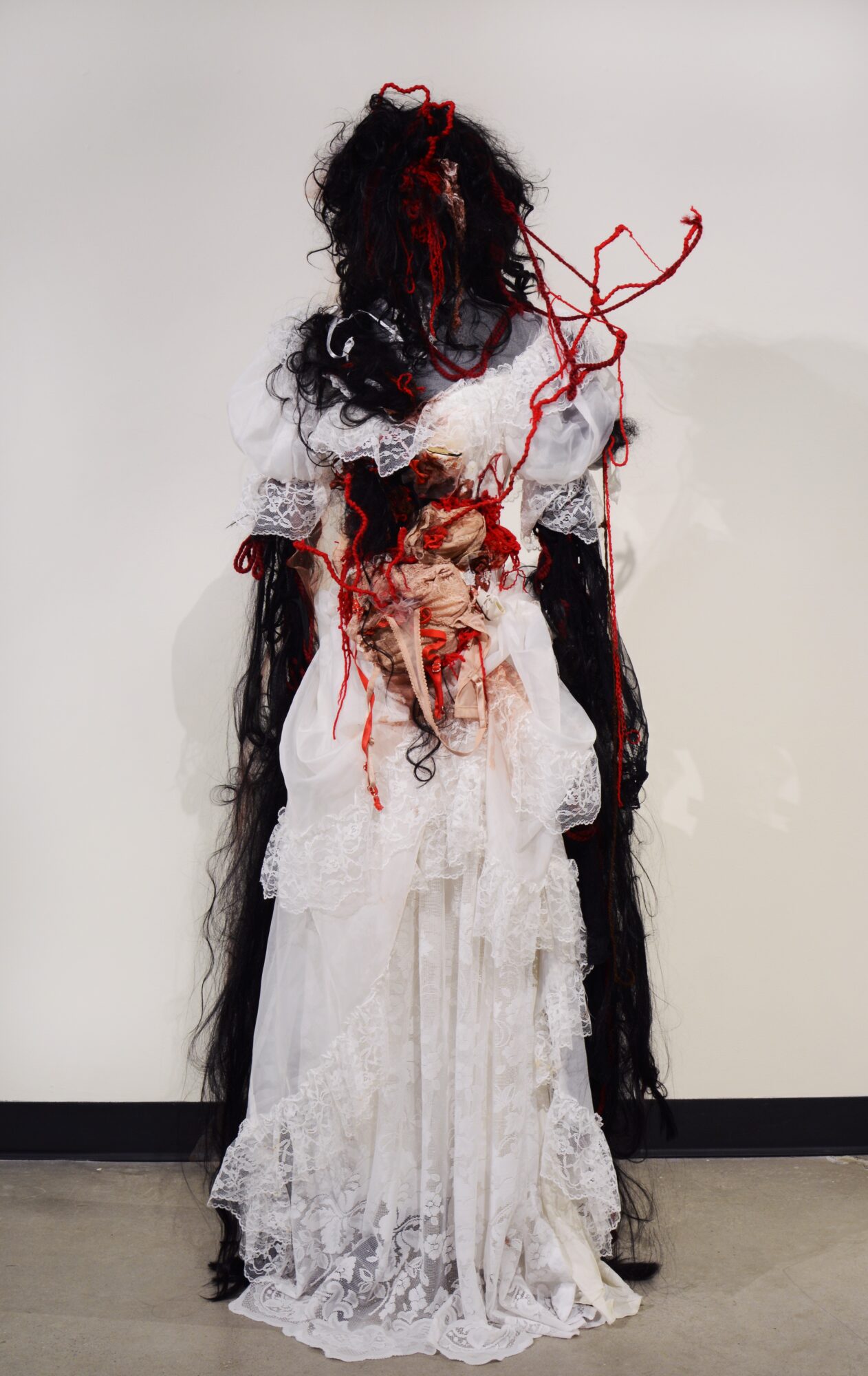 A figure in a long white lace dress with black and red hair, standing against a plain white background.