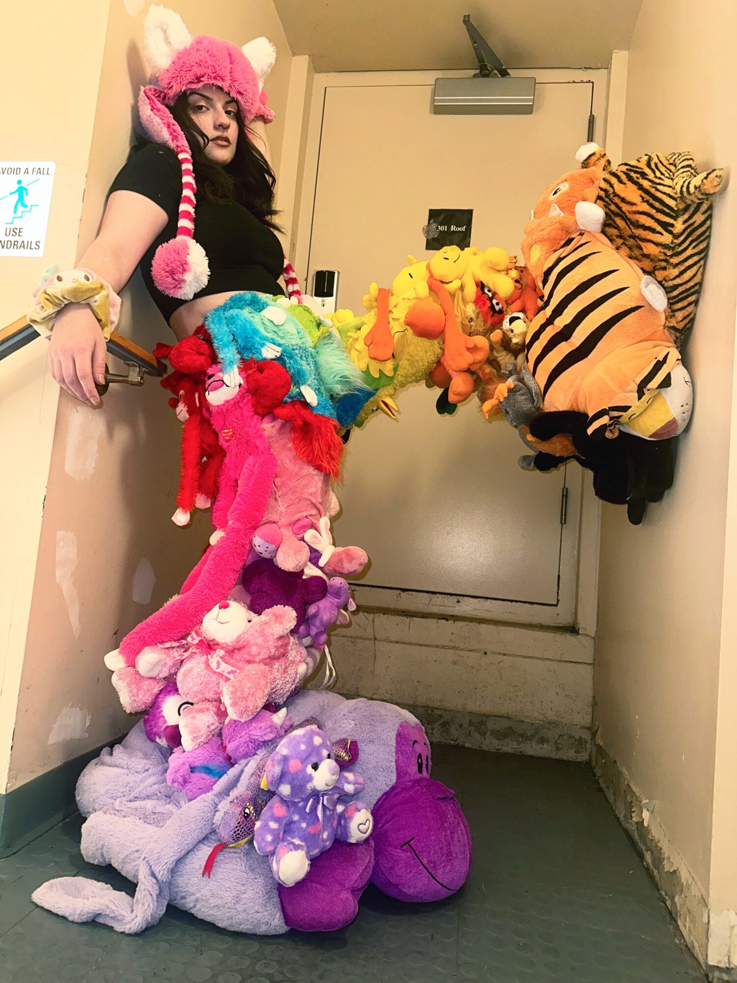 Person wearing a pink hat with animal ears and holding colorful plush toys in a narrow hallway.