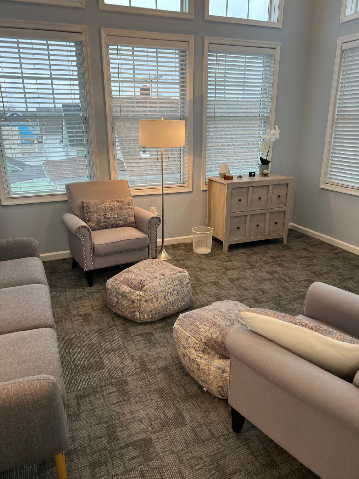 Living room with large windows, armchair, floor lamp, cabinet, and two poufs on carpeted floor.