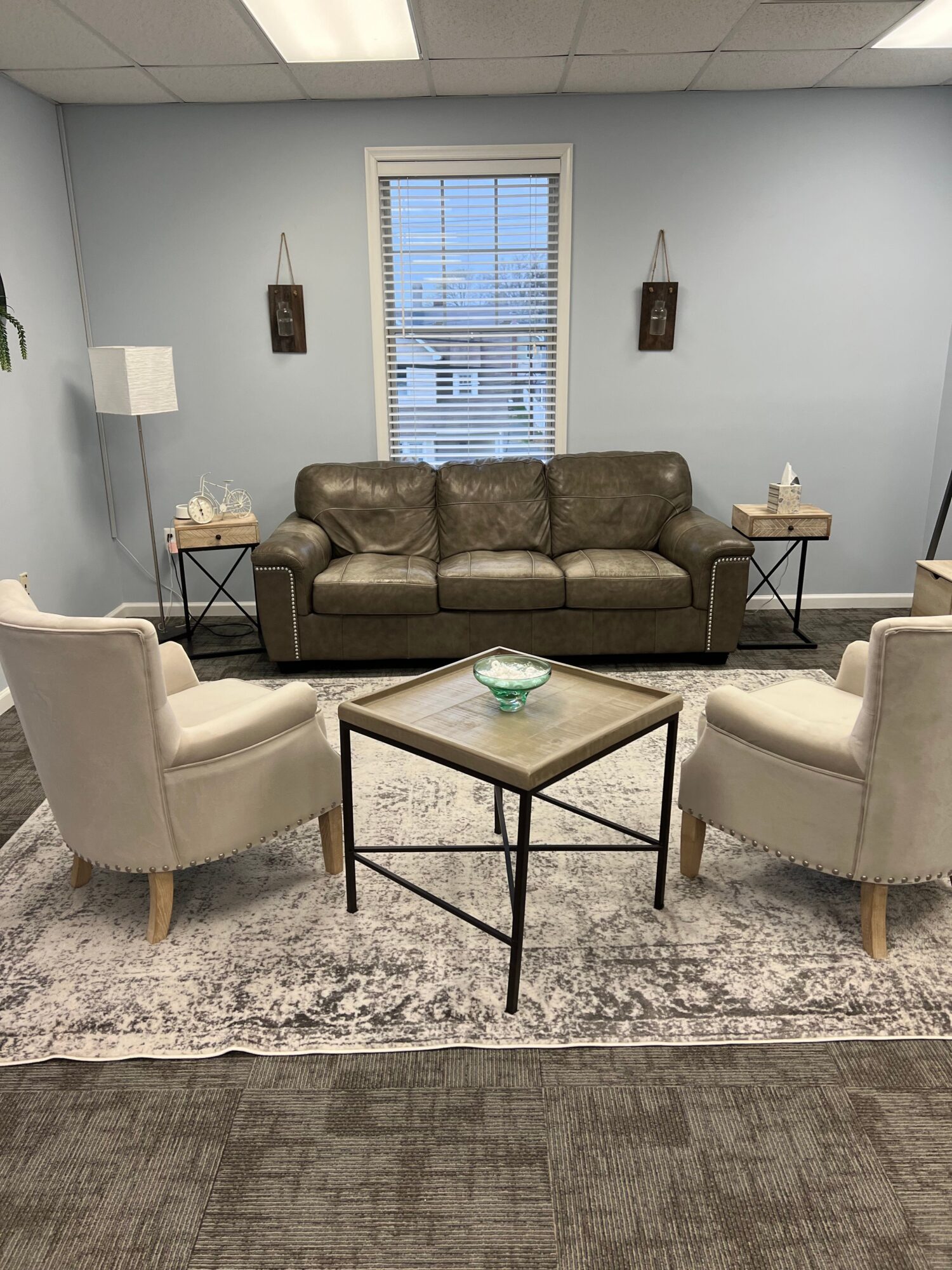 Living room with a brown sofa, two white armchairs, a glass coffee table, and a window with blinds.