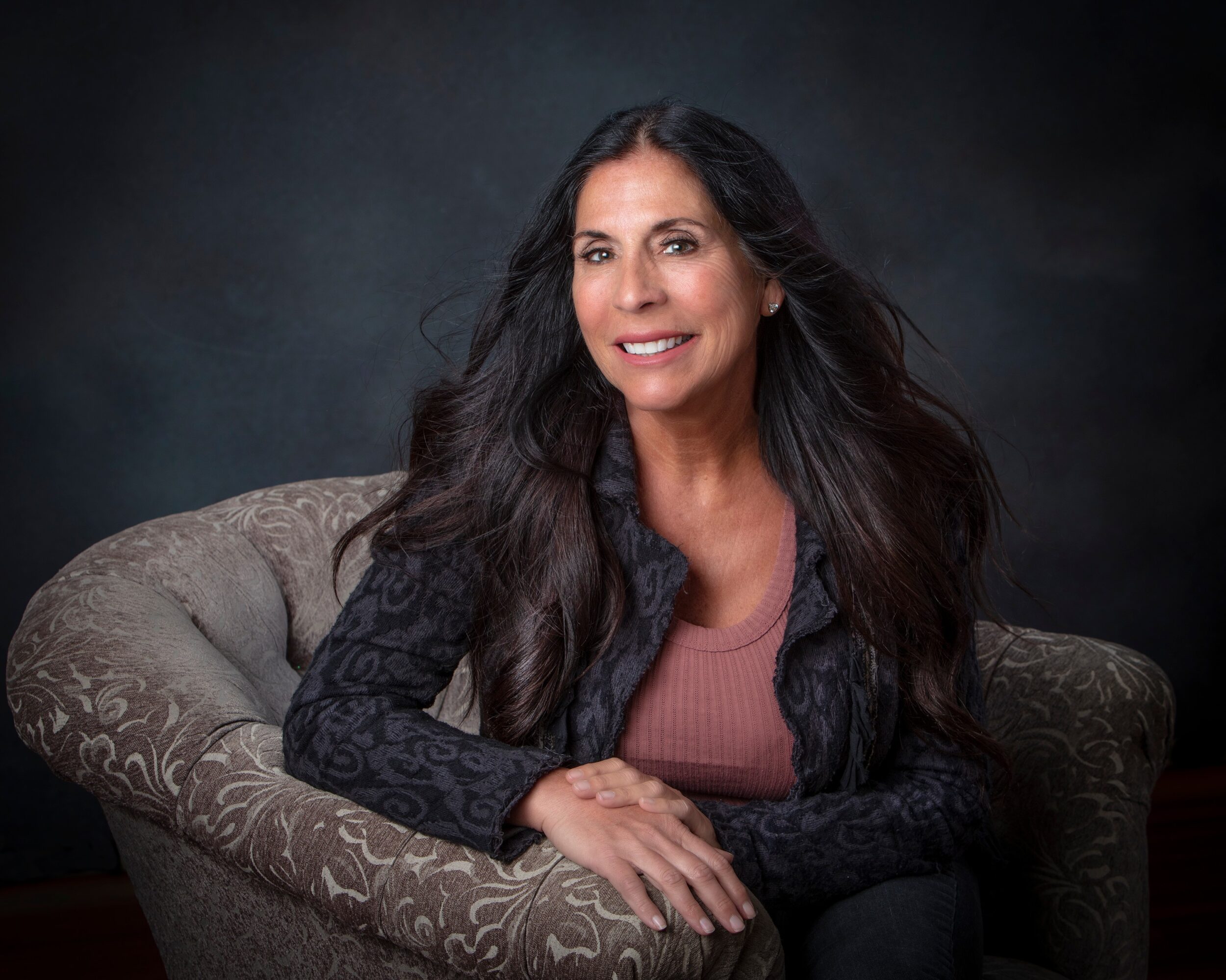 Woman with long dark hair sitting on a patterned armchair, smiling, dark background.