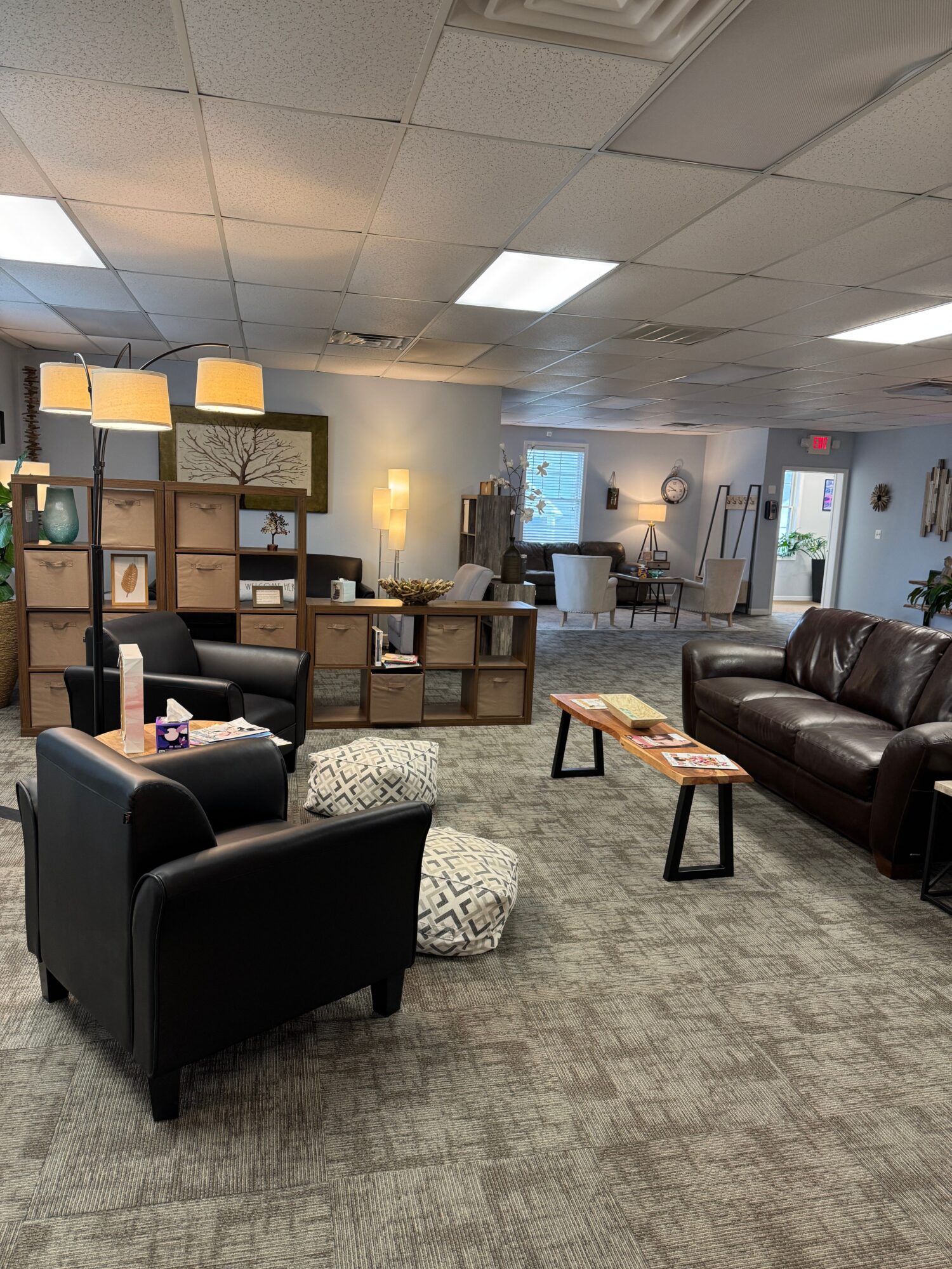 Seating area with black armchair, patterned ottomans, and two sofas in a room with shelves and lamps.