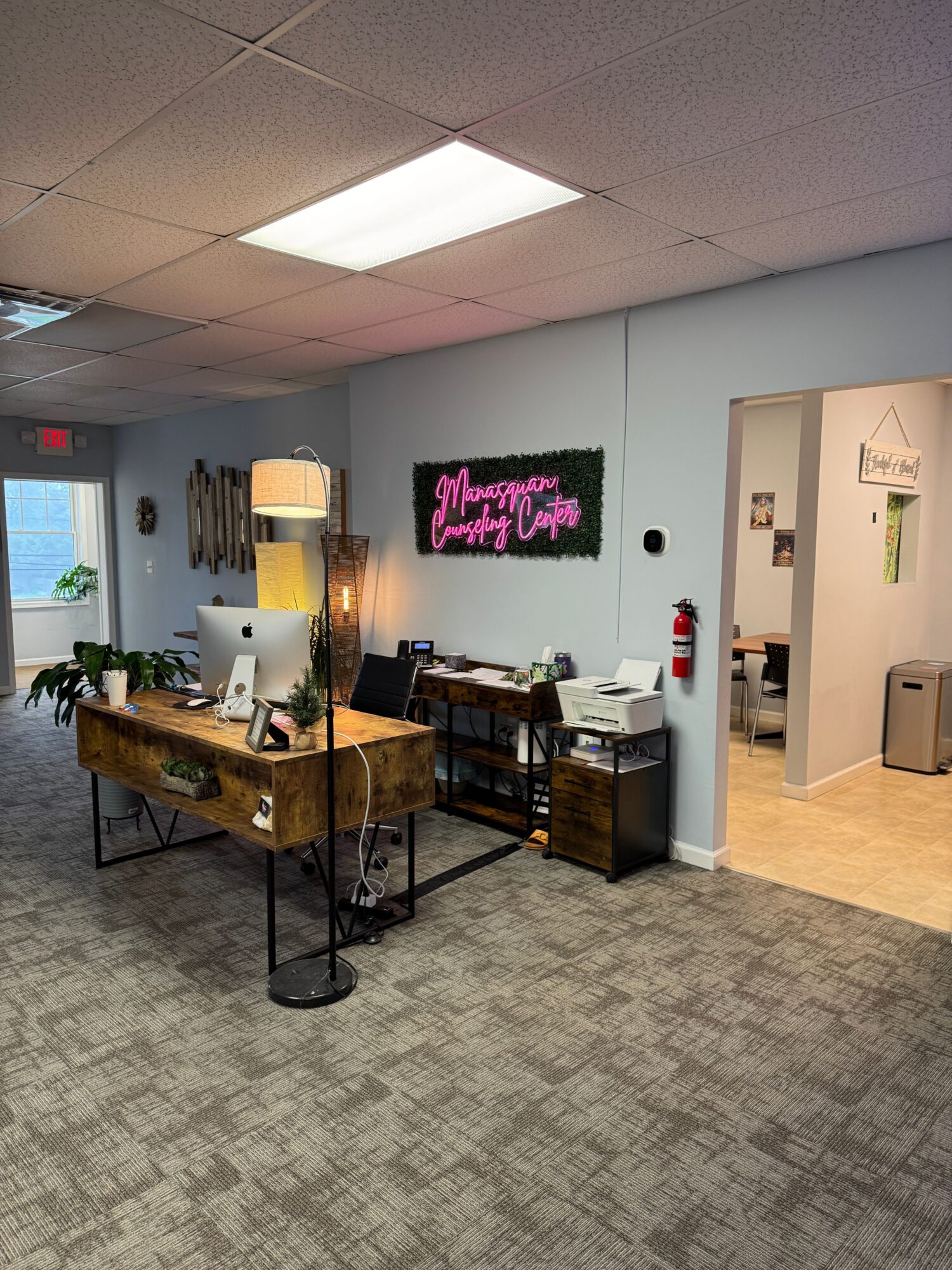 Office space with desk, computer, lamp, and wall decorations, including a neon sign and a doorway to another room.