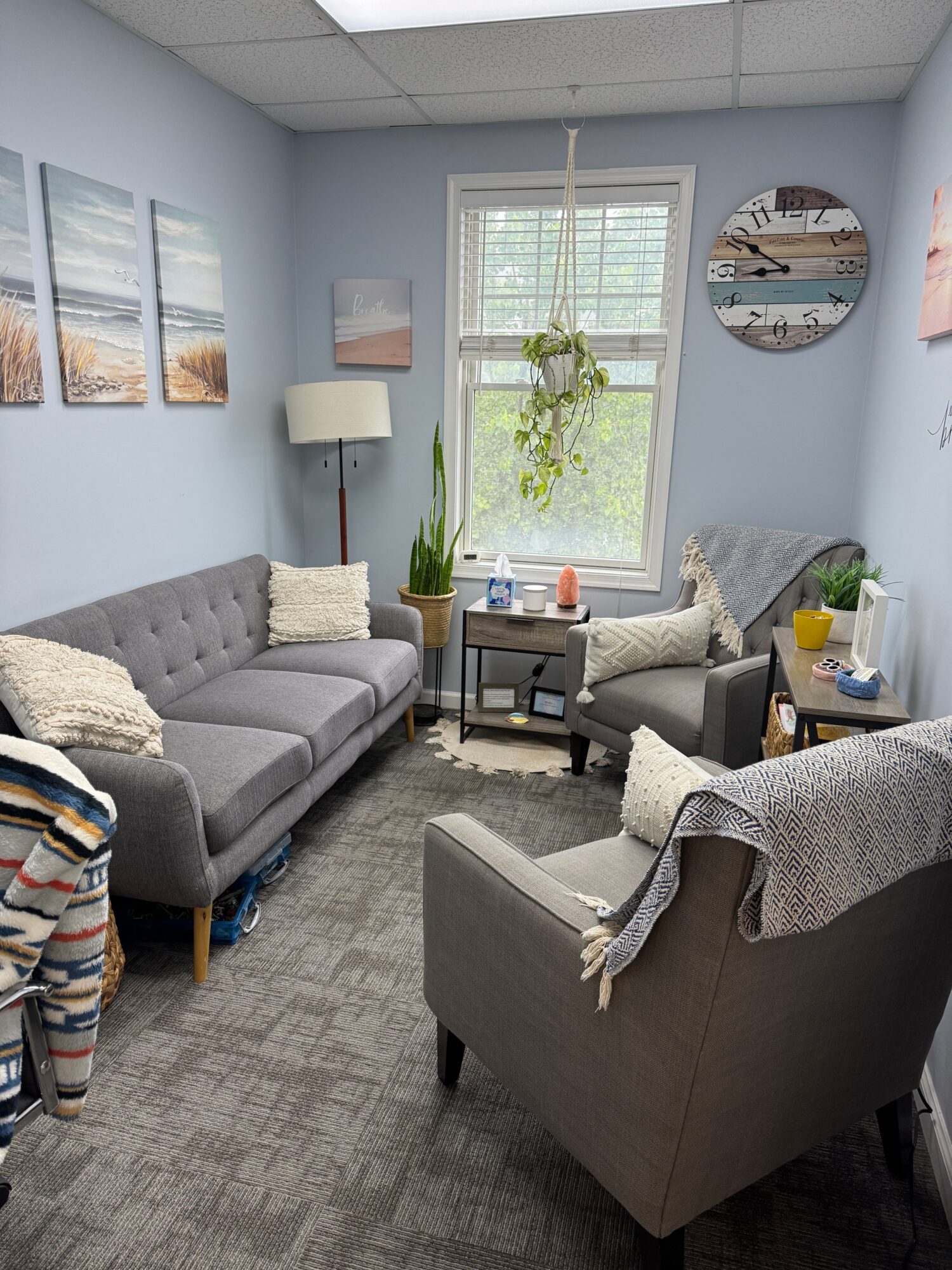Living room with gray sofa, armchair, window with blinds, wall clock, and various decorative items.