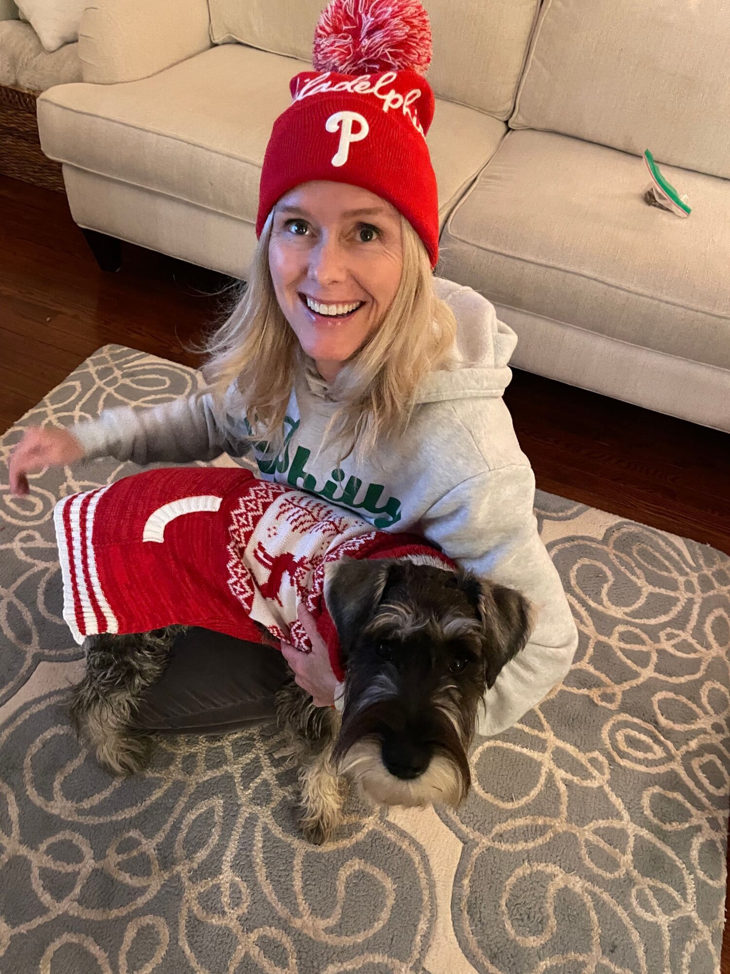 Woman wearing a red winter hat and gray hoodie, smiling with a black and gray dog in a festive sweater.