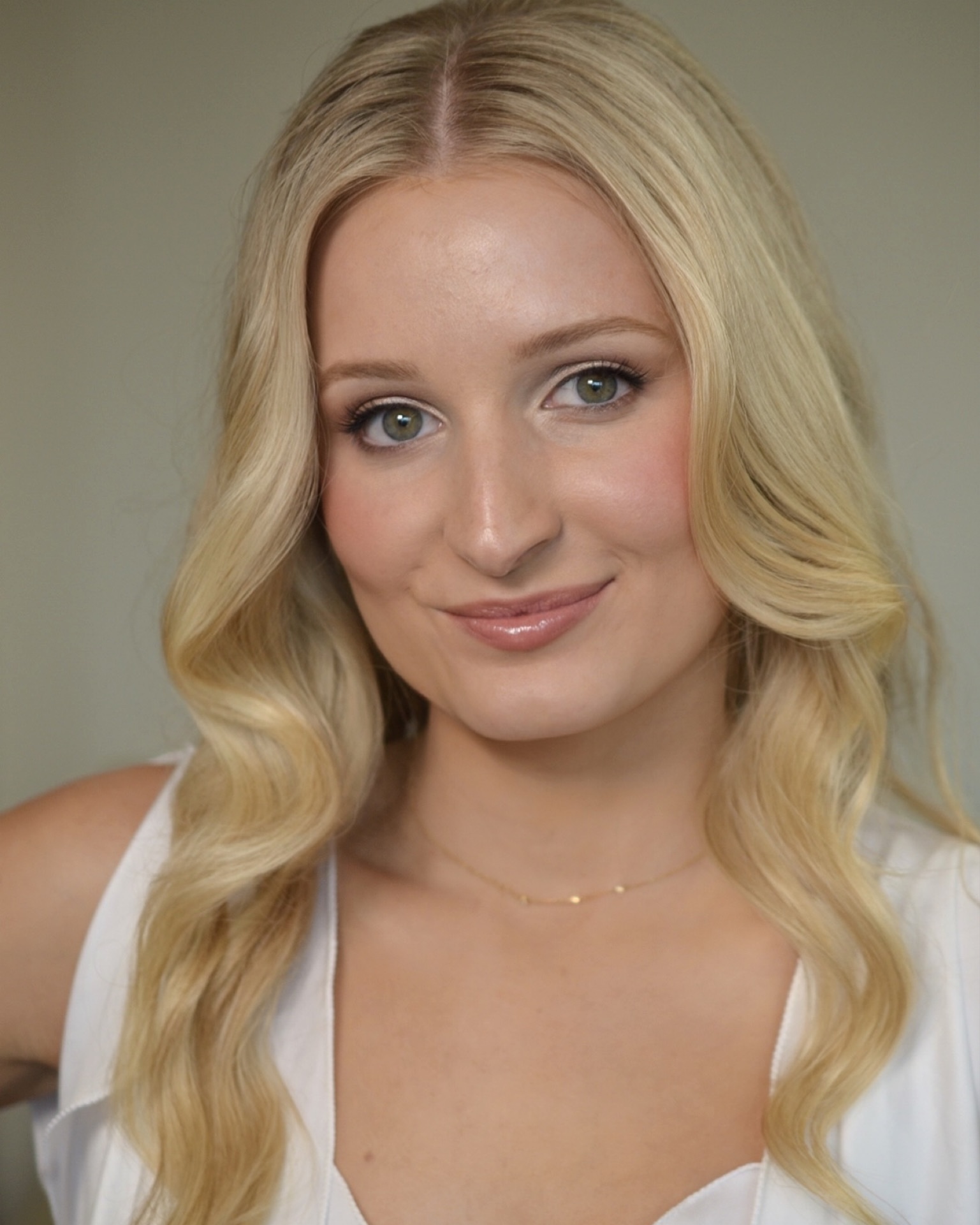 Young woman with long blonde hair, blue eyes, and a gentle smile, wearing a white top and a delicate necklace.