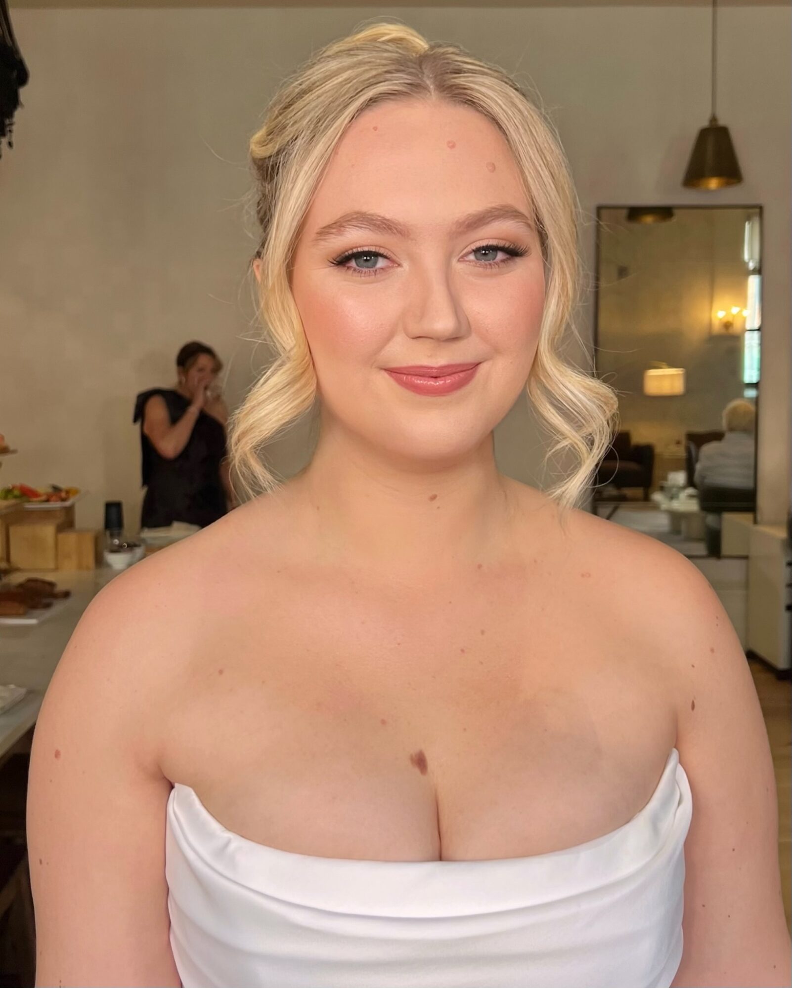 Young woman with blonde hair styled in loose waves, wearing a strapless white dress, smiling softly.