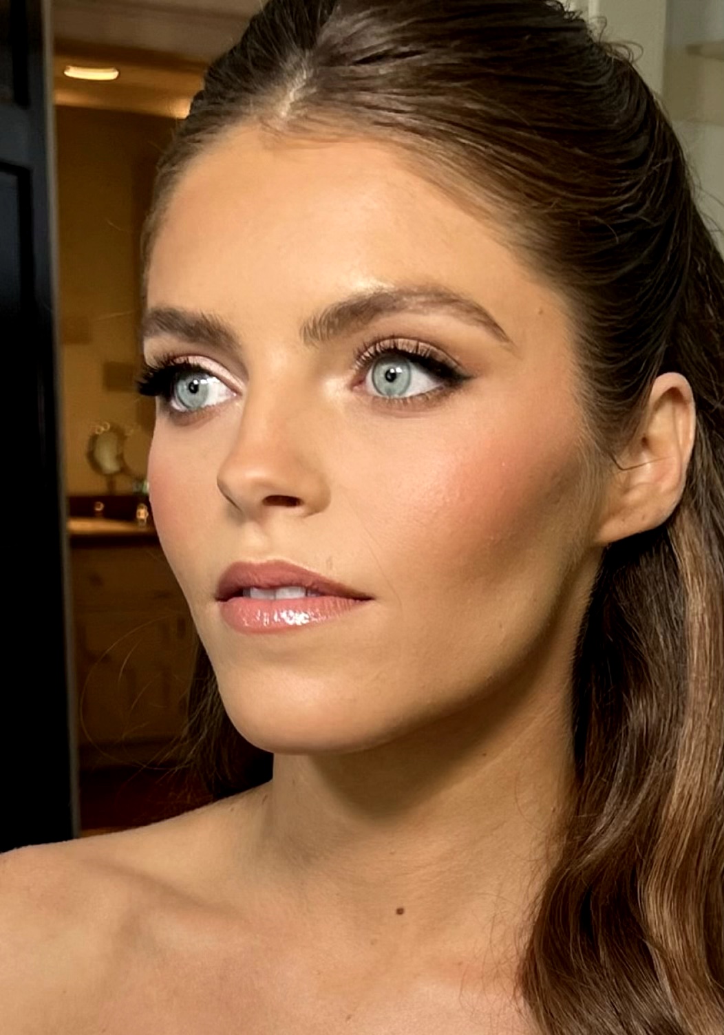 Close-up of a woman with blue eyes, brown hair, and makeup, looking to the side indoors.