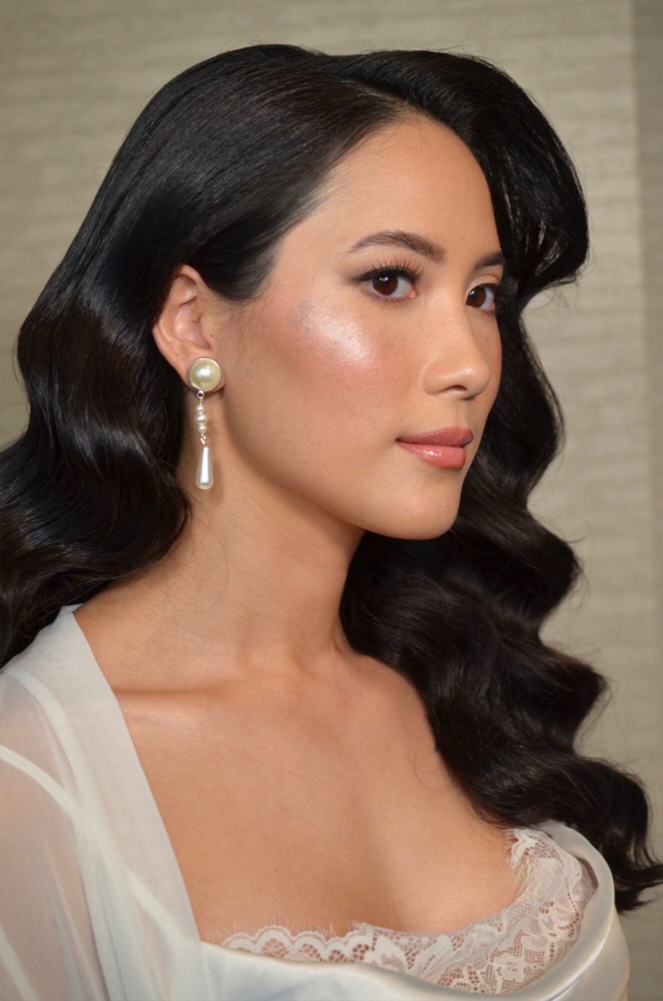 Woman with long dark wavy hair, wearing pearl earrings, looking to the side, with makeup and a white lace top.