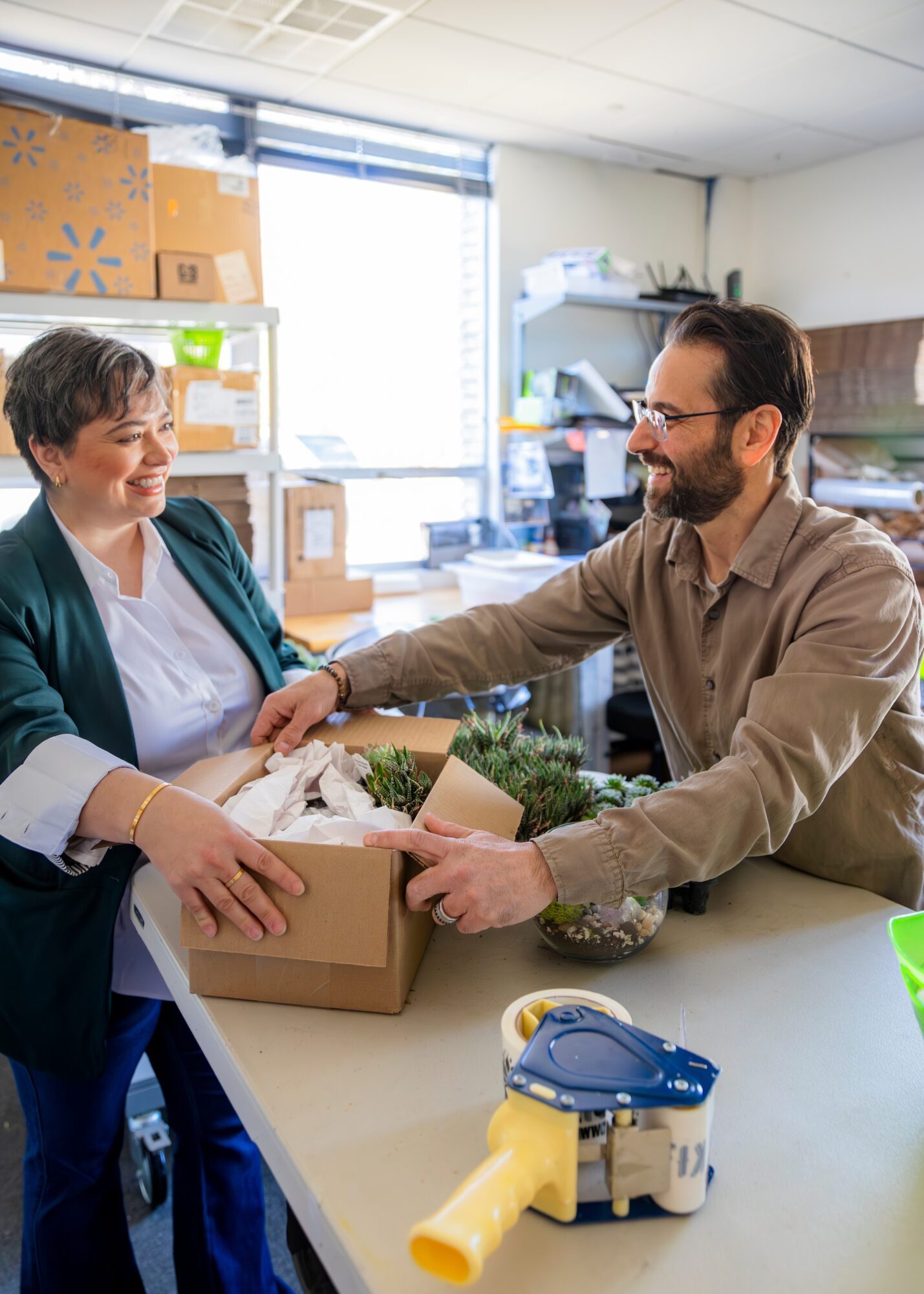 Two people exchanging a box with plants in an office, smiling and engaging in conversation.