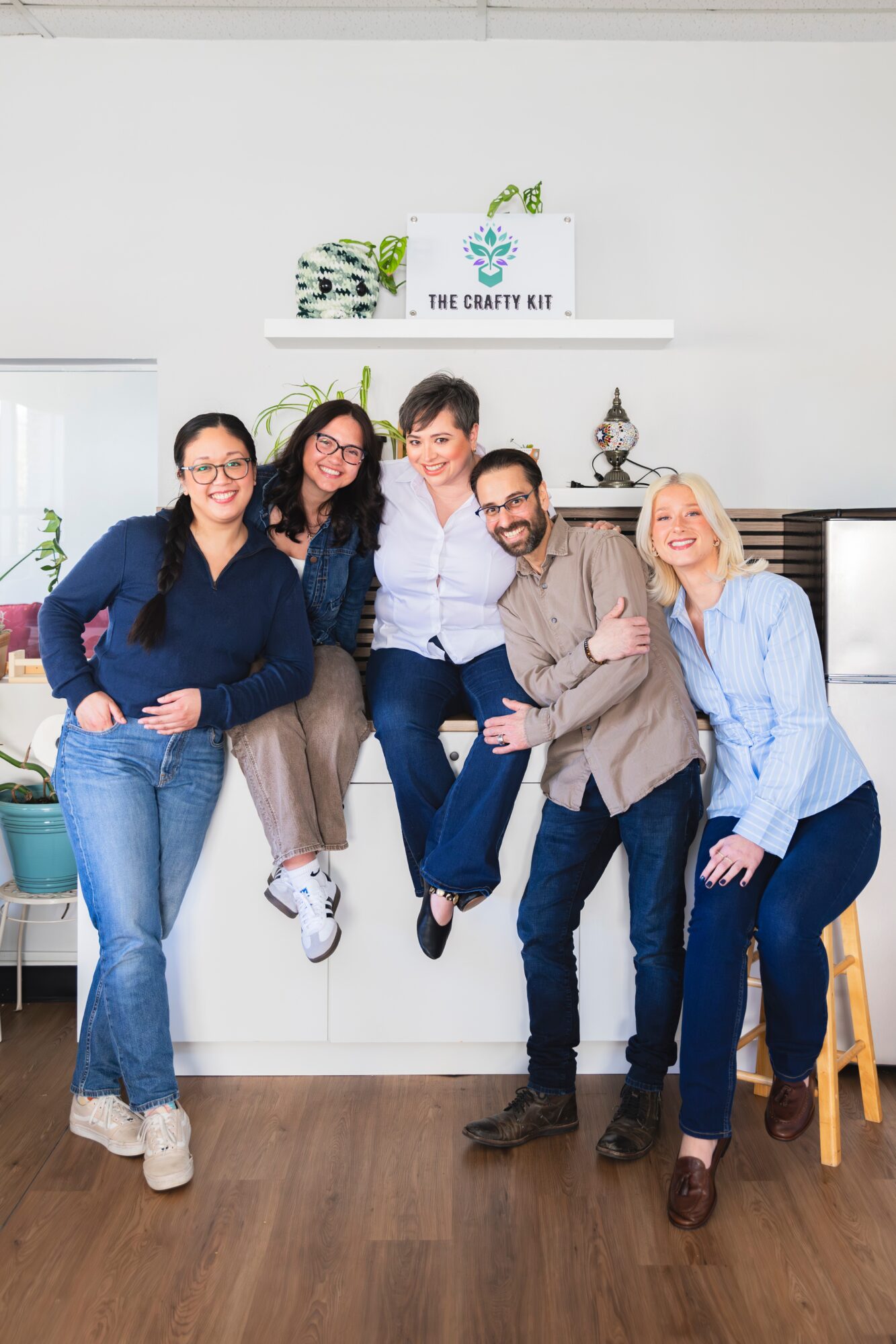 Group of five people smiling and posing indoors, with a white wall and a sign reading 'The Crafty Kit' behind them.