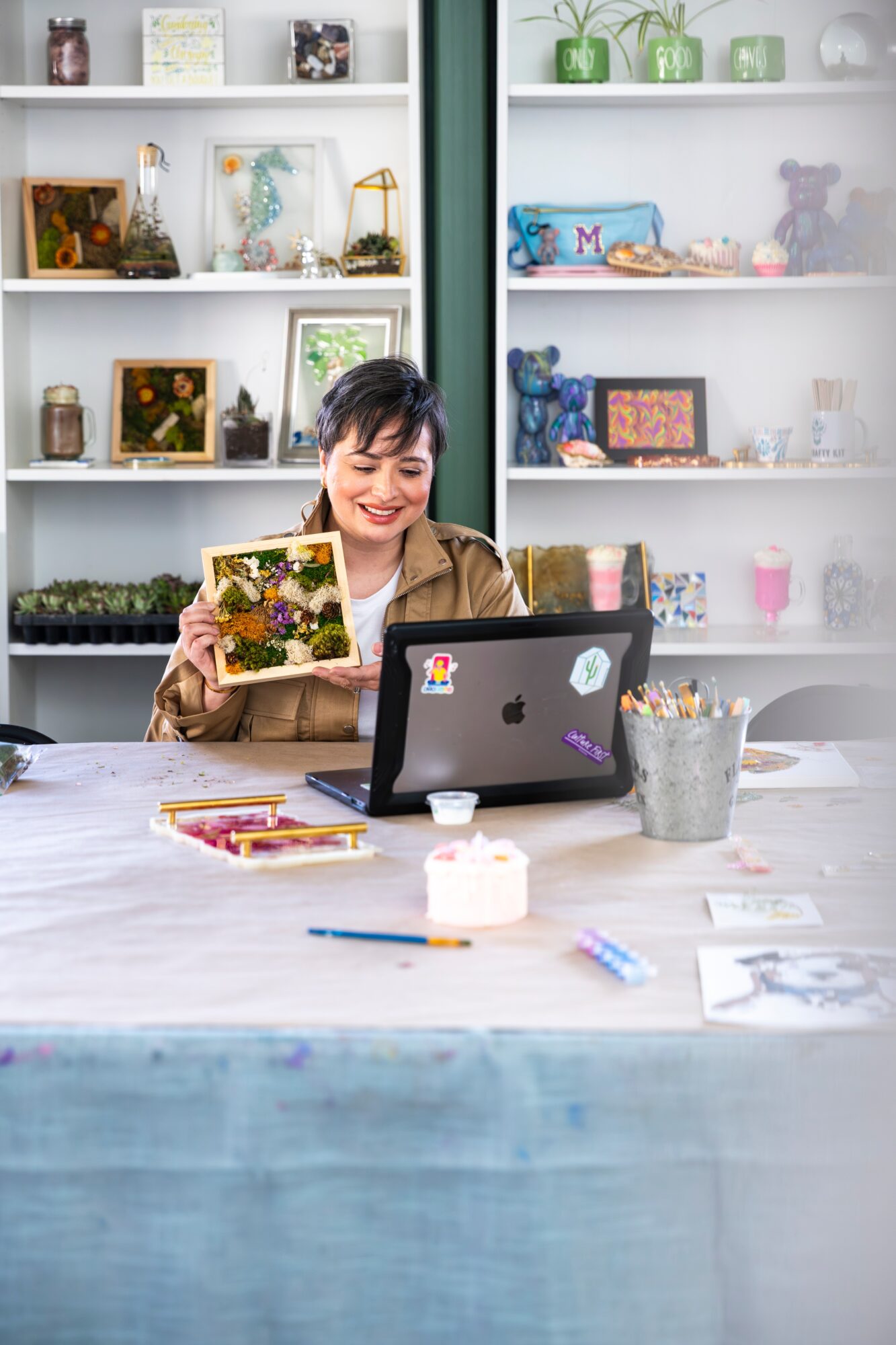 Woman smiling at a laptop with art supplies on a table in a room with shelves filled with decorative items.