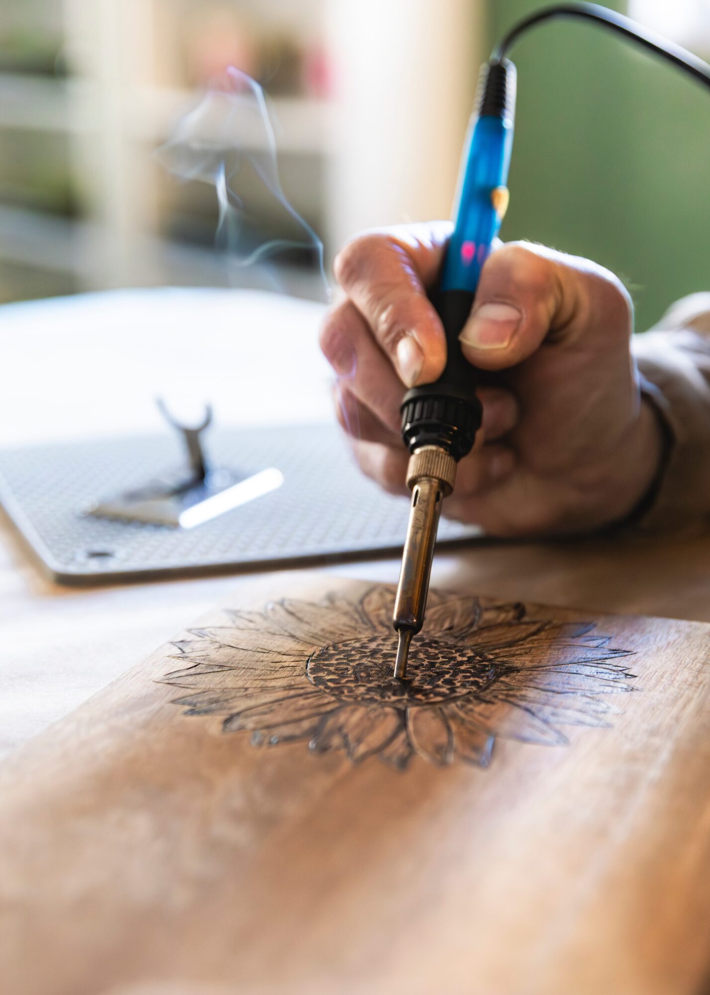 Person using a soldering iron on a wooden surface with a blurred background, tools nearby, and smoke rising.