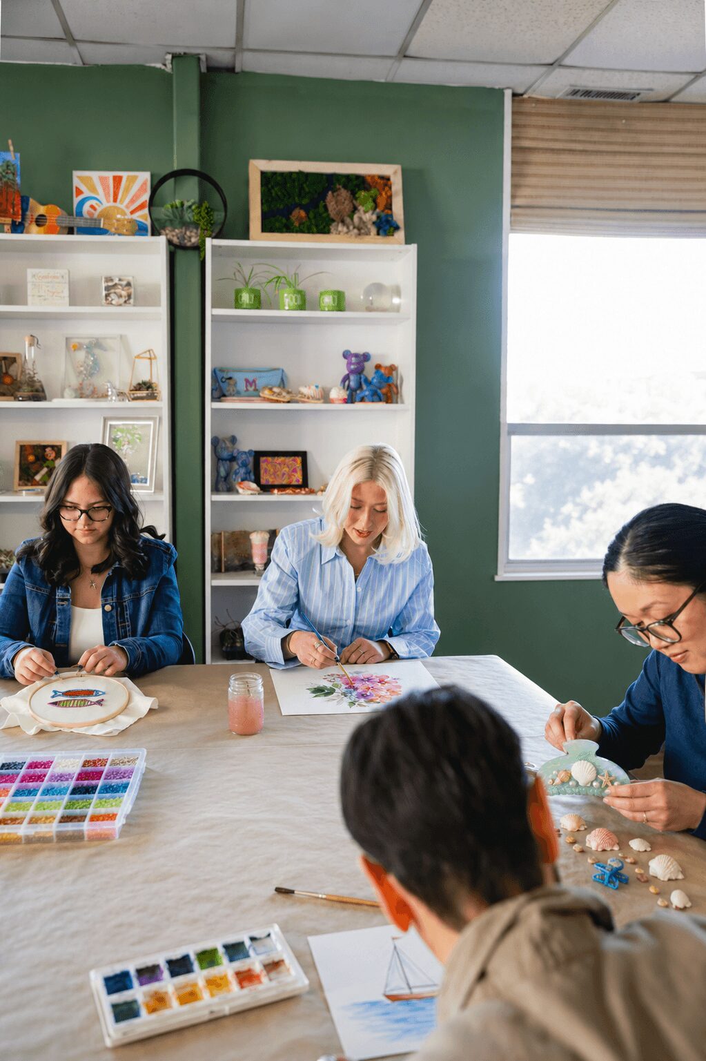 Four women sit around a table in a room with green walls and shelves, working on arts and crafts projects.