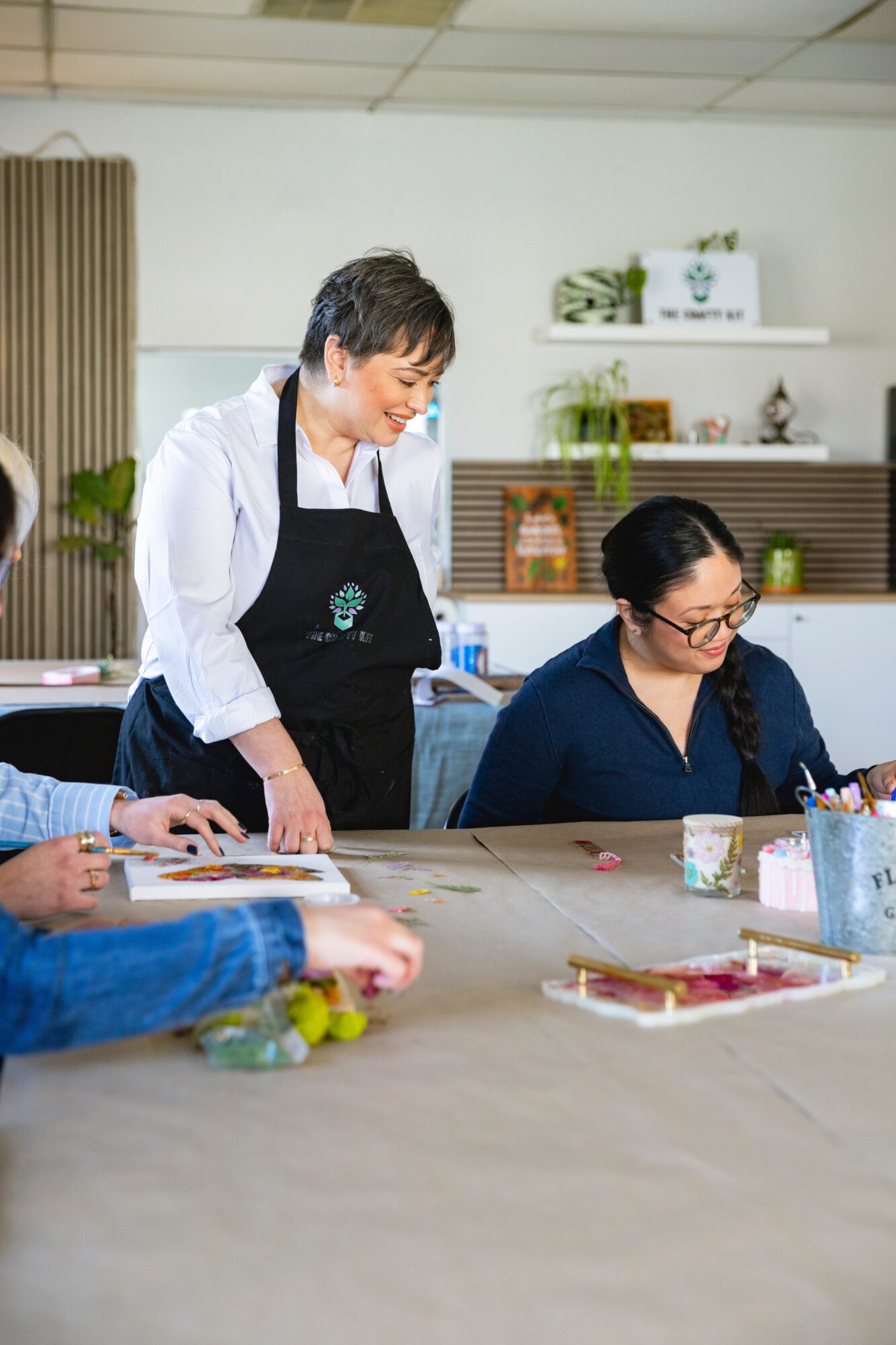 Two women in a kitchen, one standing and smiling, the other sitting and writing, with various items on the table.