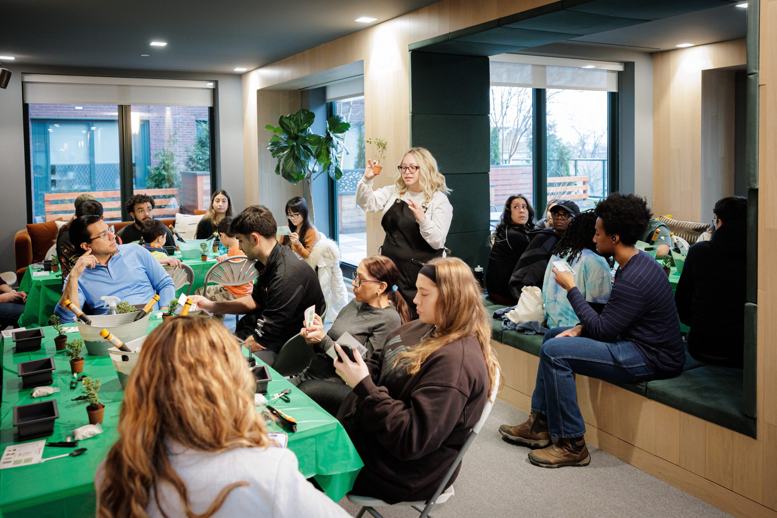 Group of people seated around a table in a modern room with large windows, some using laptops and phones.