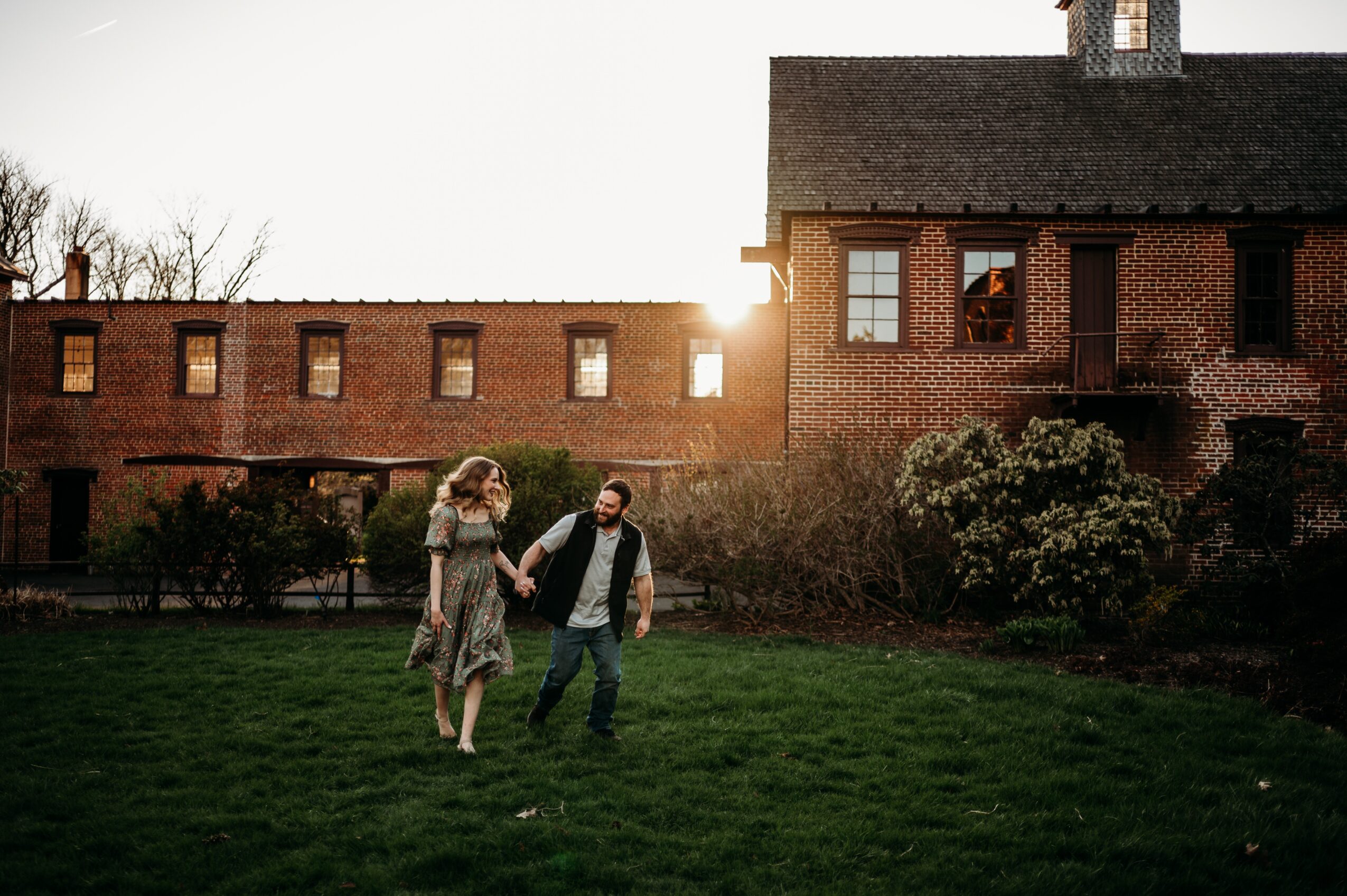Two people walking on grass near a brick building with trees and bushes, sunlight in the background.