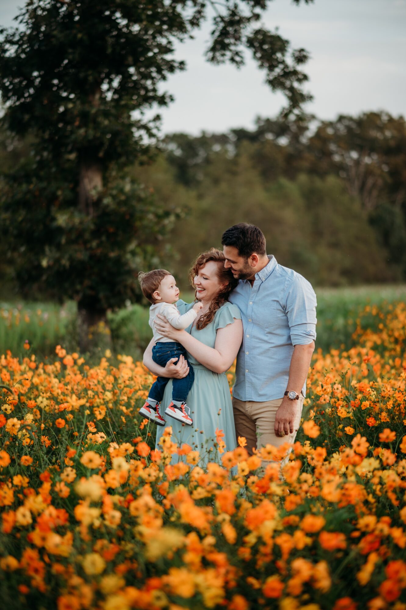 Family of three standing in a field of orange flowers with trees in the background, smiling and interacting.