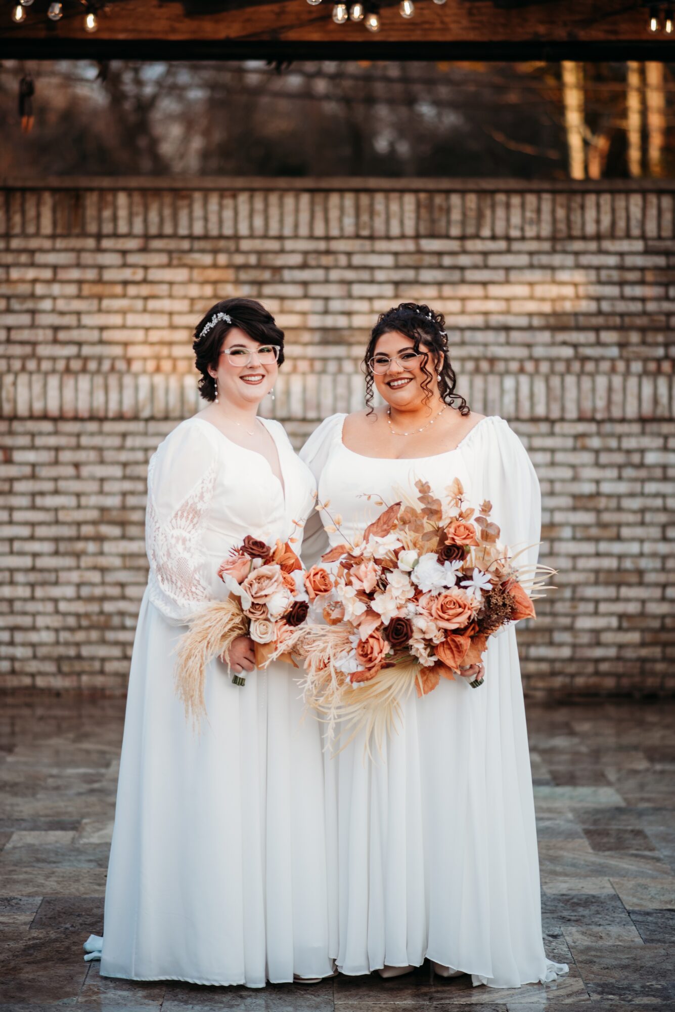 Two women in white dresses holding a large bouquet of flowers, standing outdoors in front of a brick wall.
