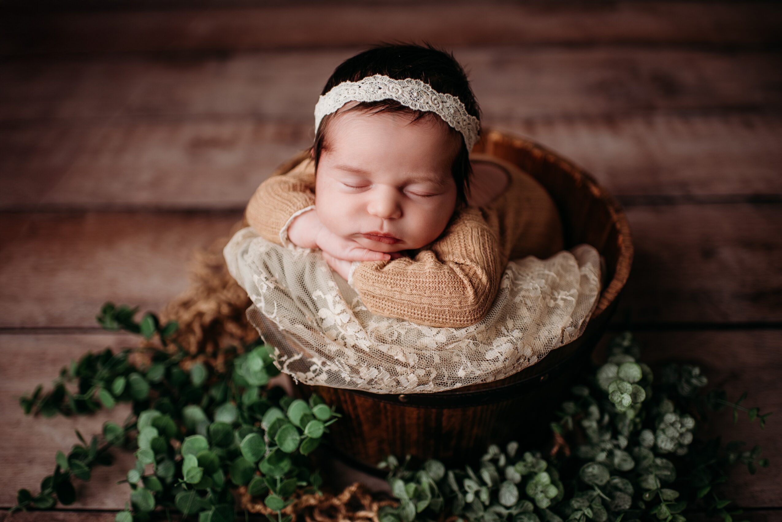 Baby with head resting on hands, wearing a headband, in a basket, surrounded by green plants, on wooden surface.