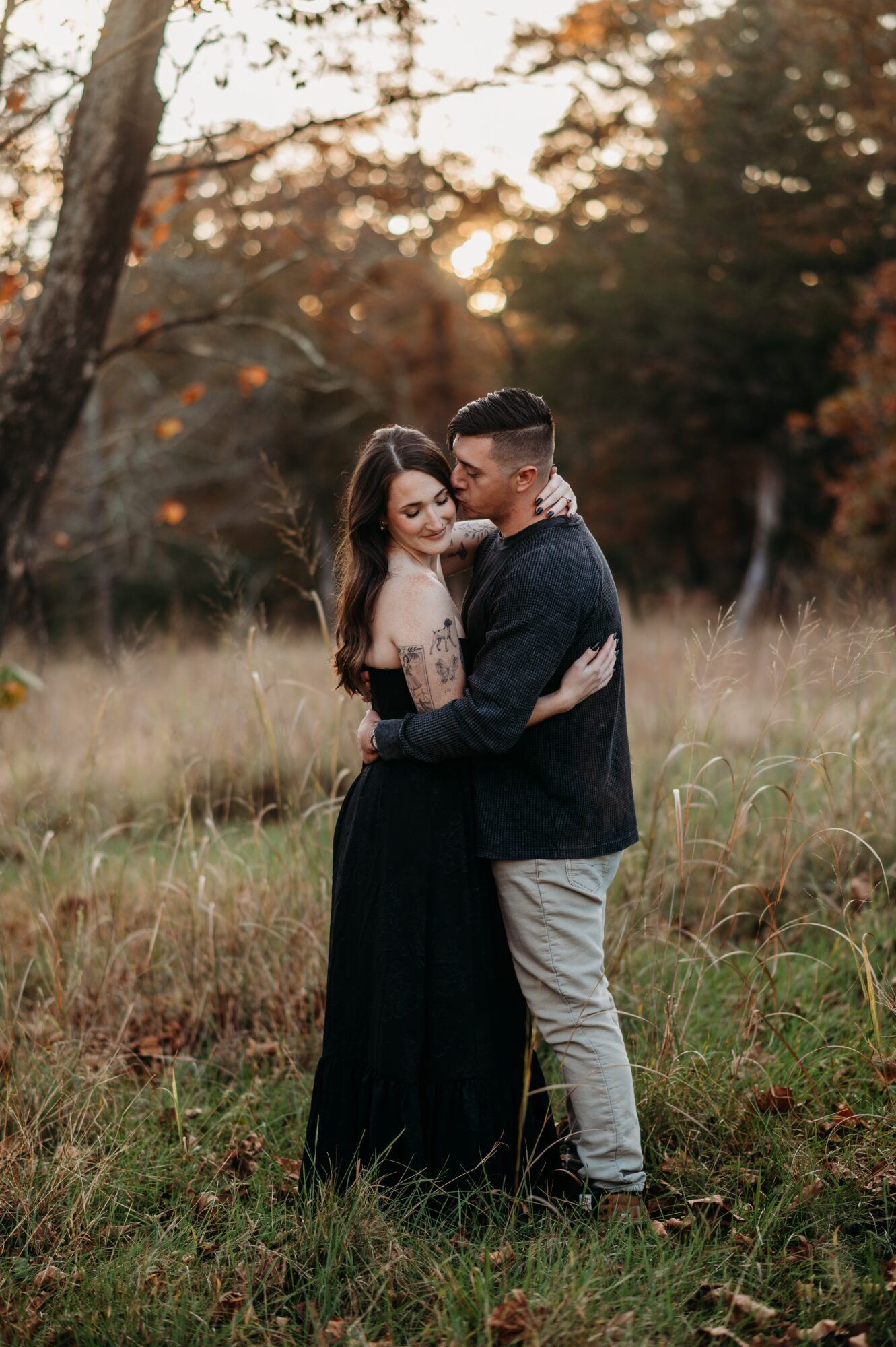 A couple stands embracing in a grassy field with trees and sunset in the background.