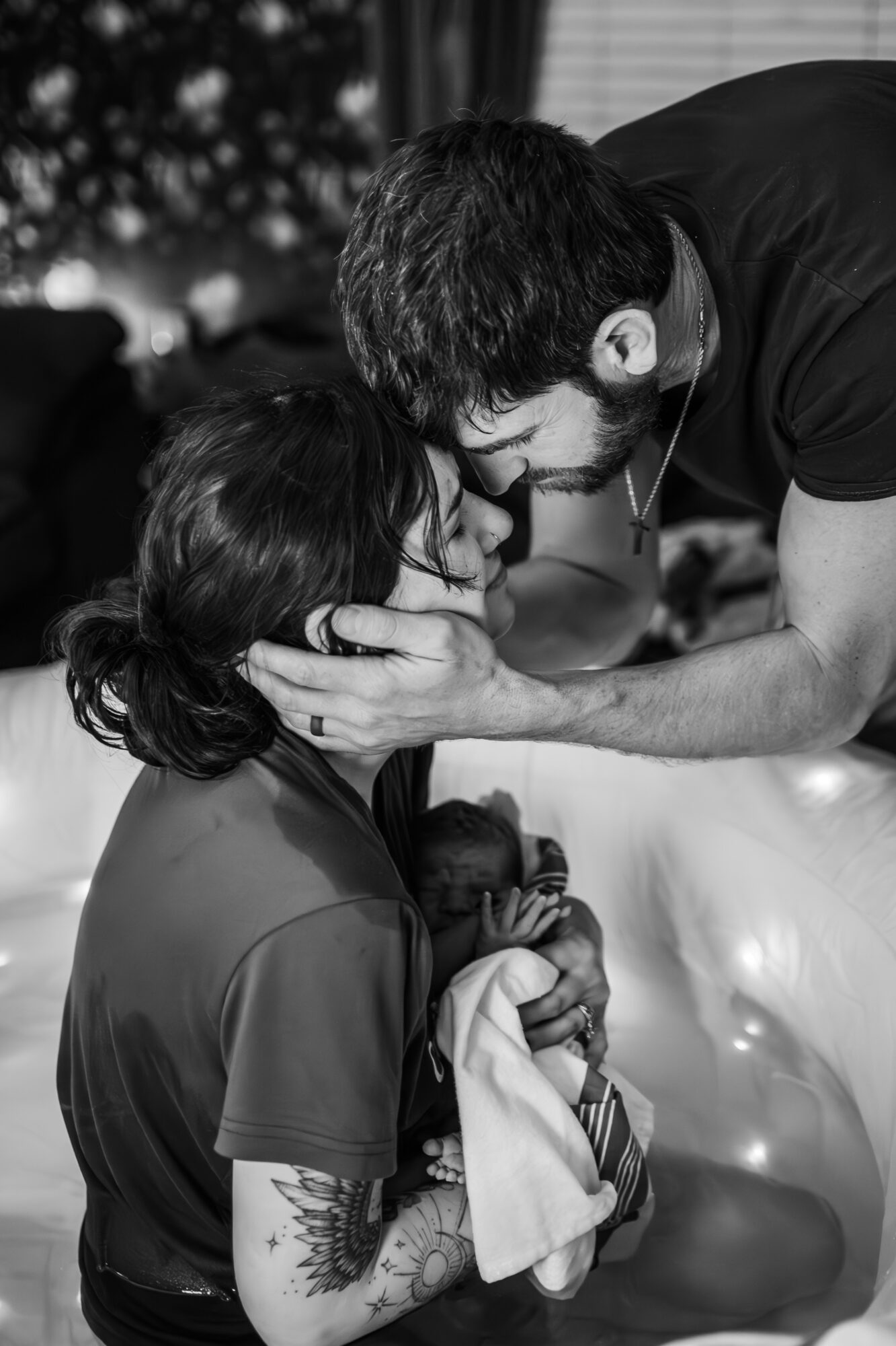 Man and woman with eyes closed, touching foreheads, holding a newborn baby, in a close, intimate moment, black and white.